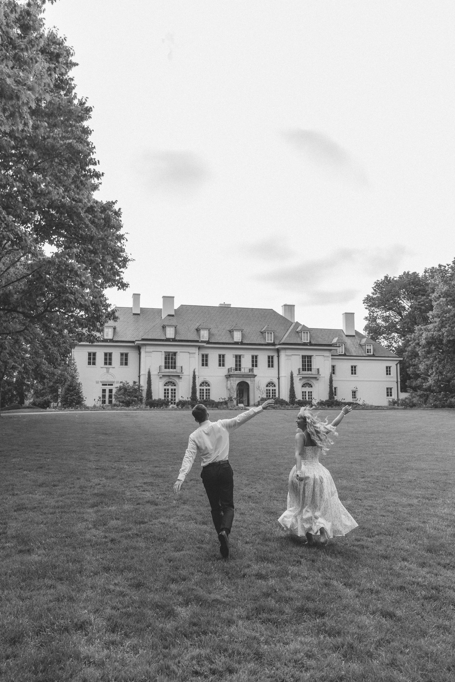 Black and white photo of bride and groom running toward a grand estate mansion on a lawn.