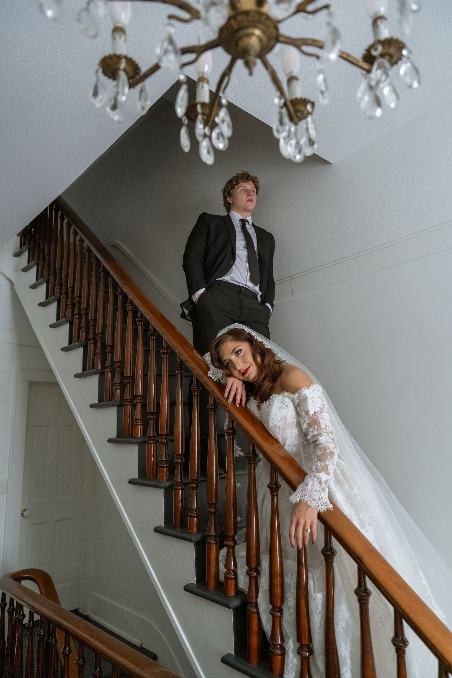Bride and groom posing dramatically on a wooden staircase beneath a vintage crystal chandelier.
