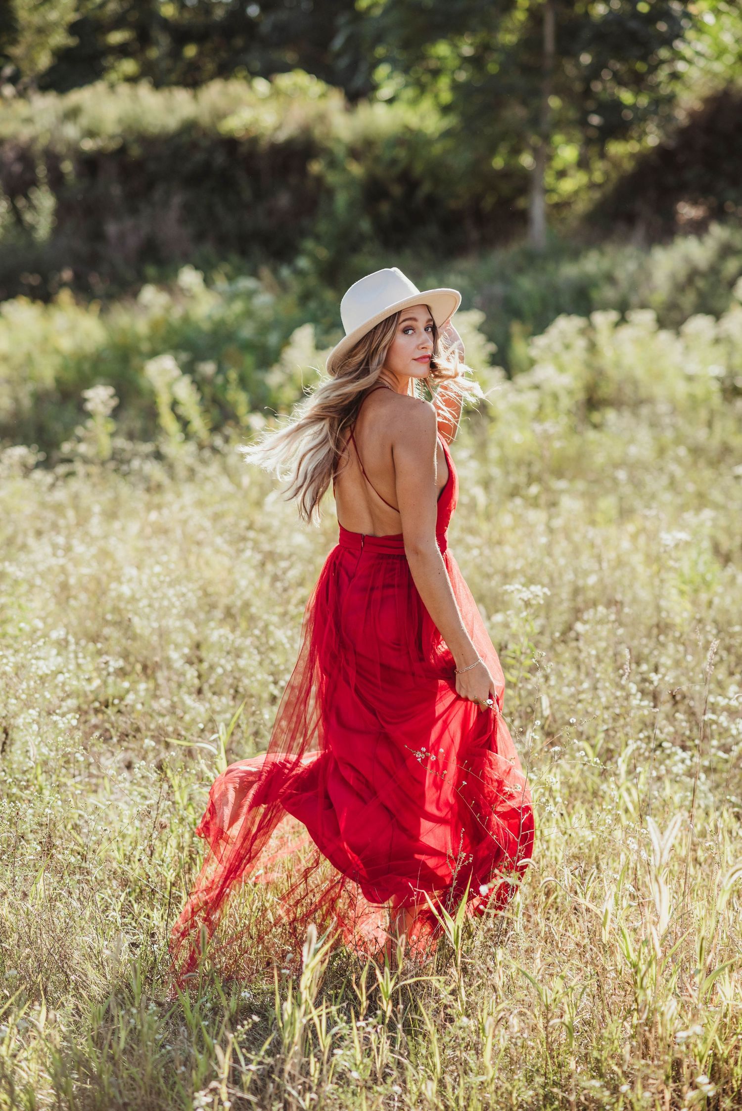 Woman in a flowing red backless dress and white hat walking through a wildflower meadow.