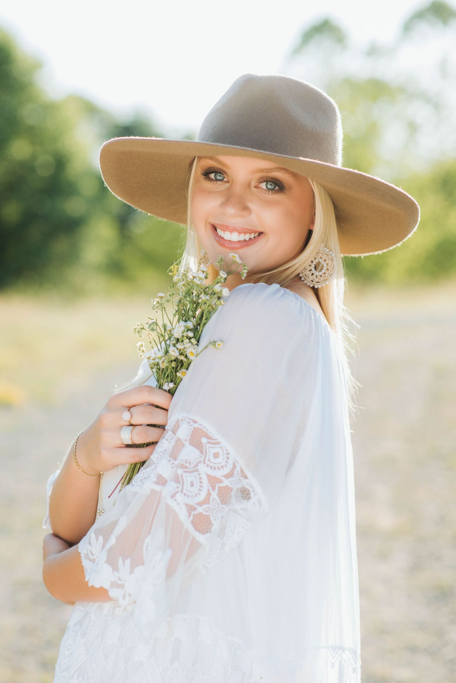 Smiling blonde woman in wide-brim hat and white lace top holding wildflowers in a sunny outdoor setting.