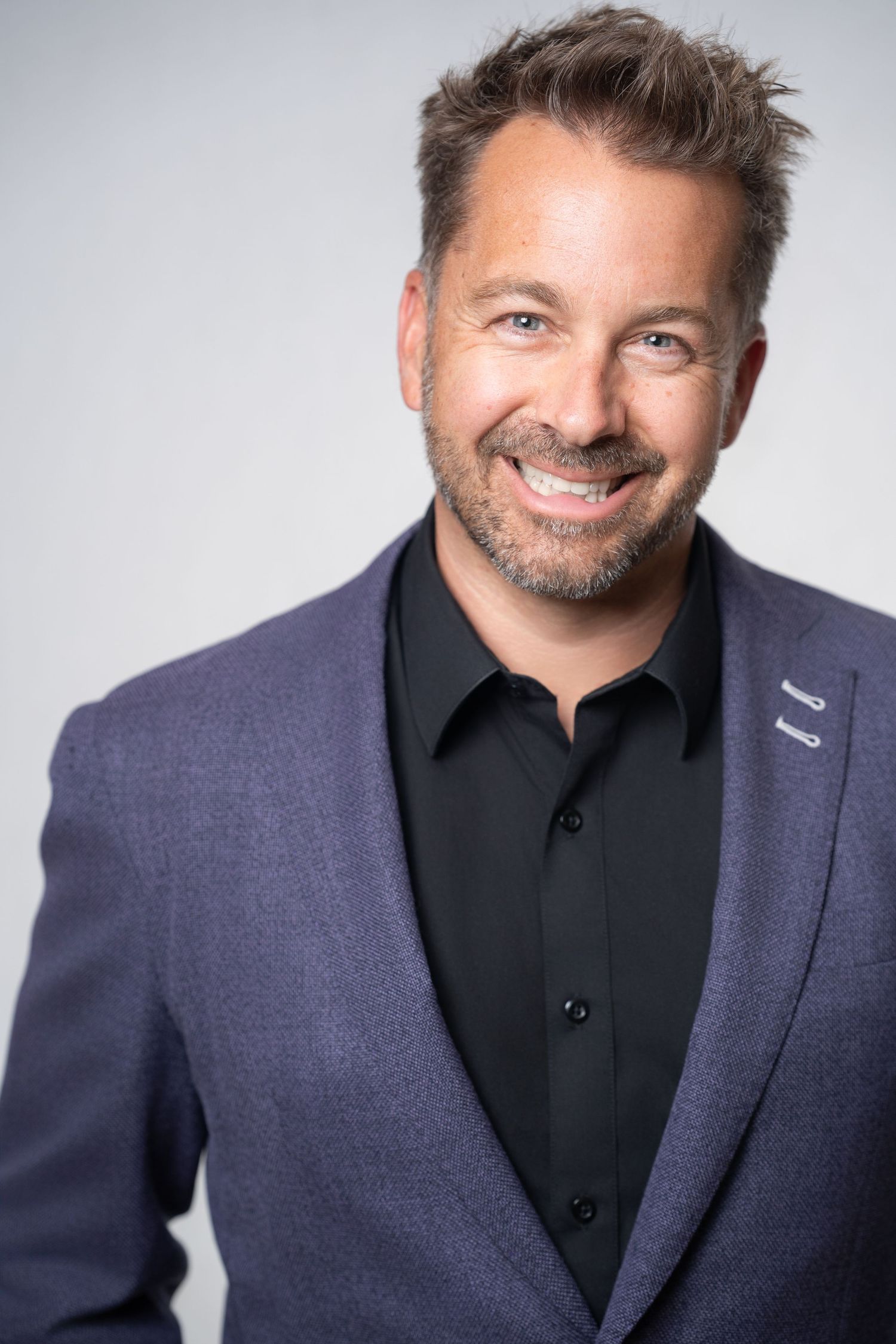 Smiling man with stubble in purple blazer and black shirt on gray background.