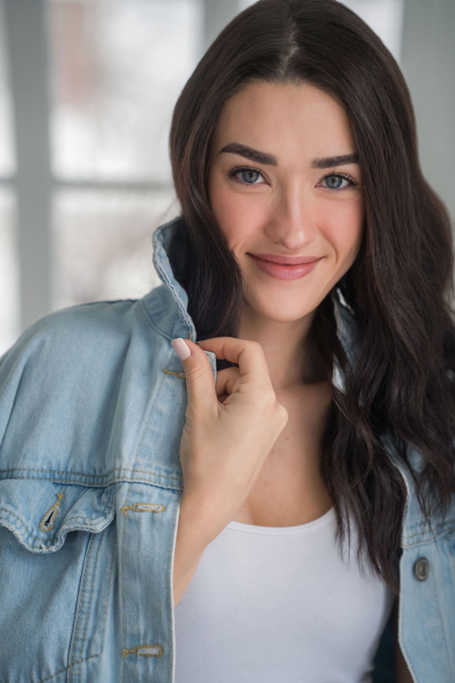 Young brunette woman in denim jacket smiling with blue eyes indoors.