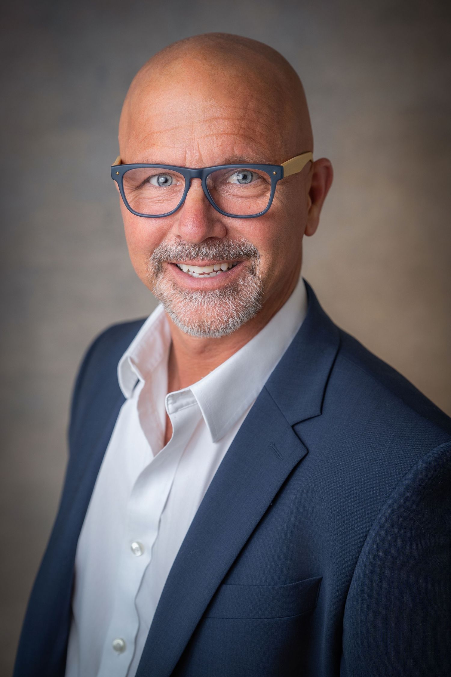 Bald man with blue glasses and gray beard smiling in navy blazer.