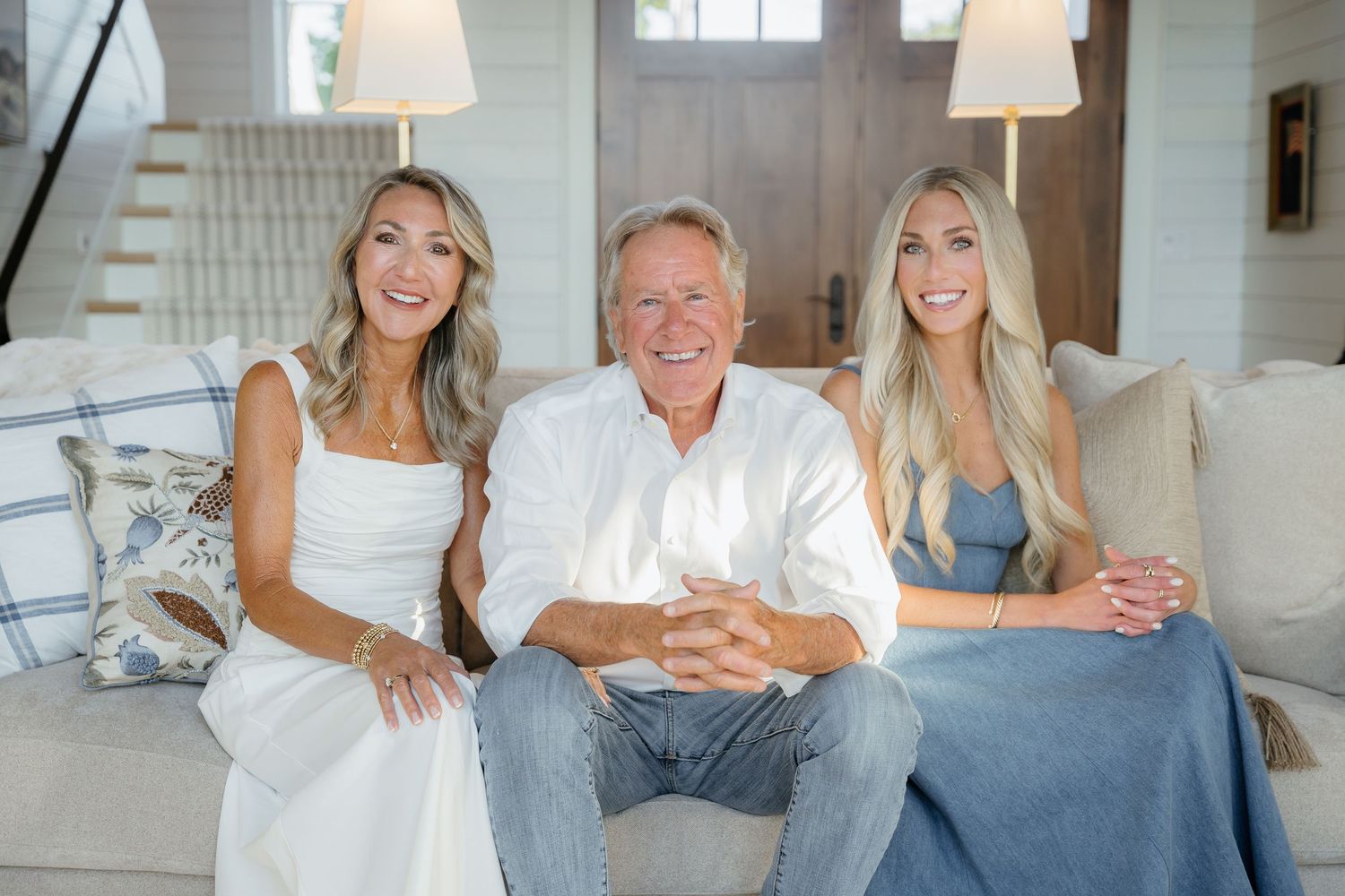 A smiling man with two blonde women sitting together on a couch in a bright modern home.