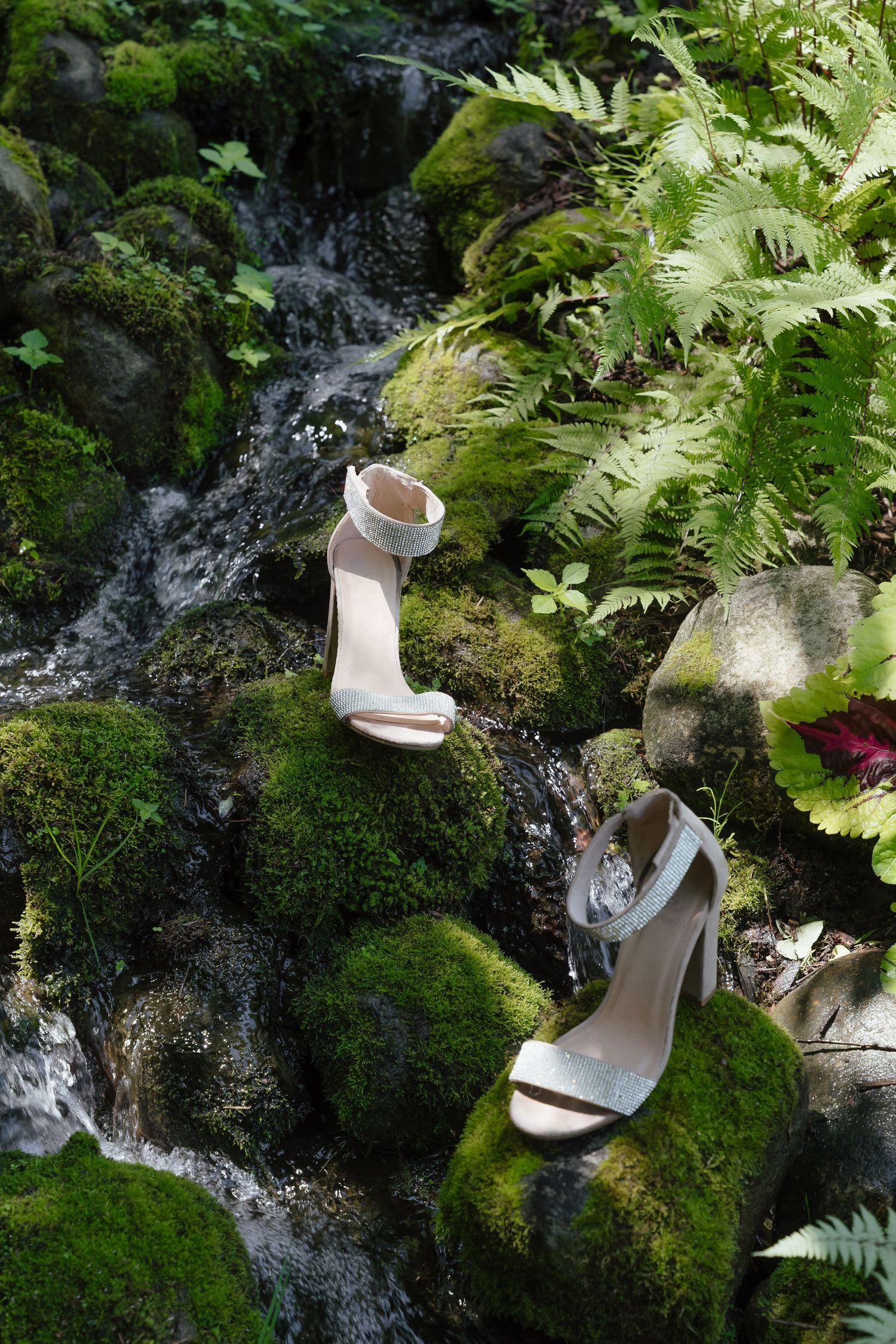 White bridal heels placed on moss-covered rocks beside a small waterfall surrounded by lush ferns.