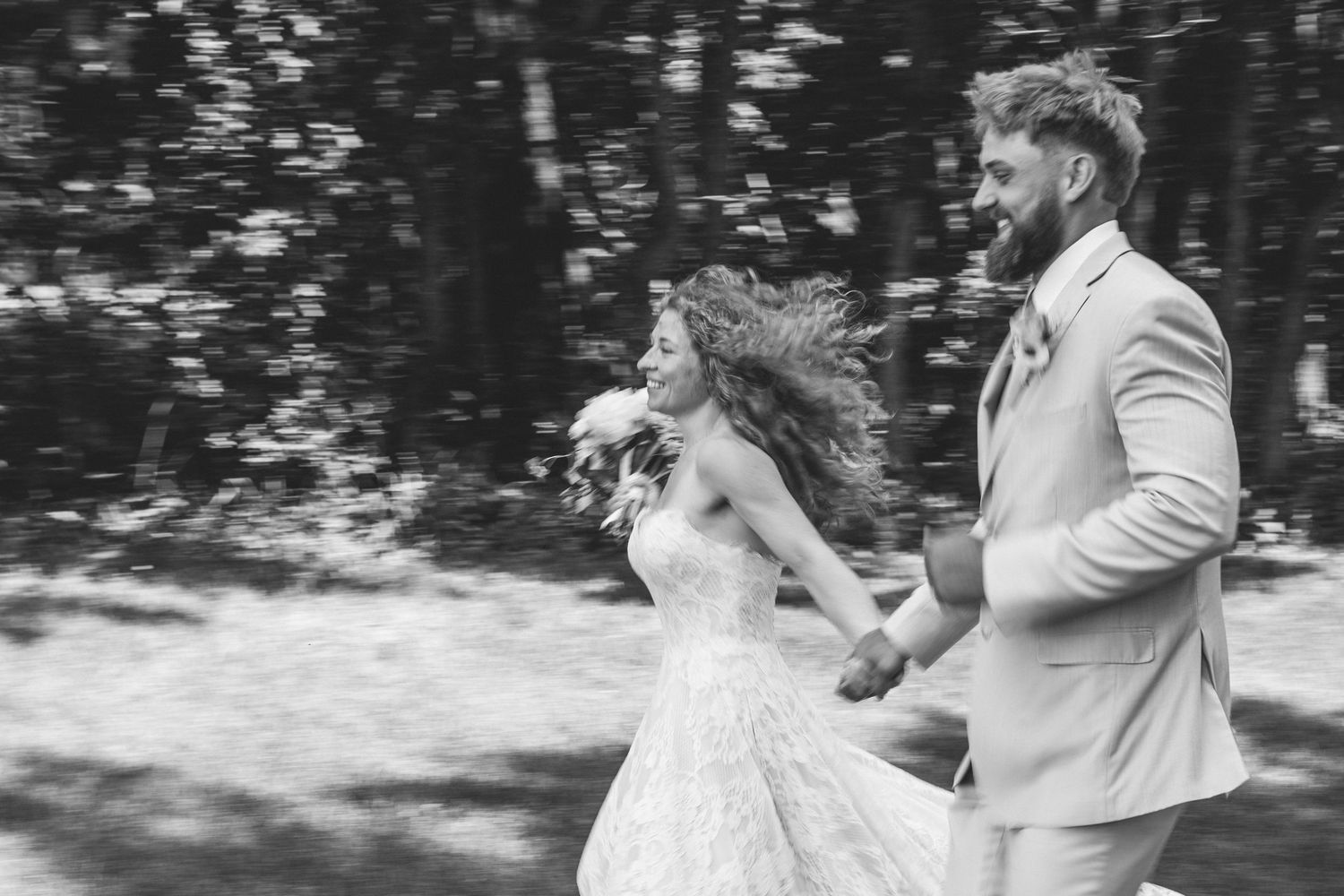 Joyful bride and groom running hand-in-hand through a sunlit forest in black and white wedding photo.
