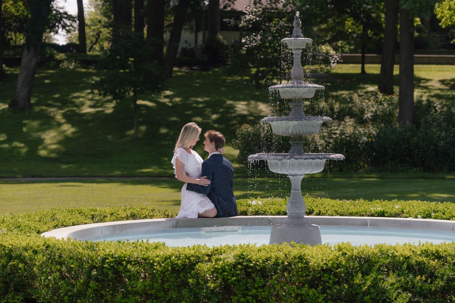 Bride and groom embracing beside ornate stone fountain in lush garden setting.