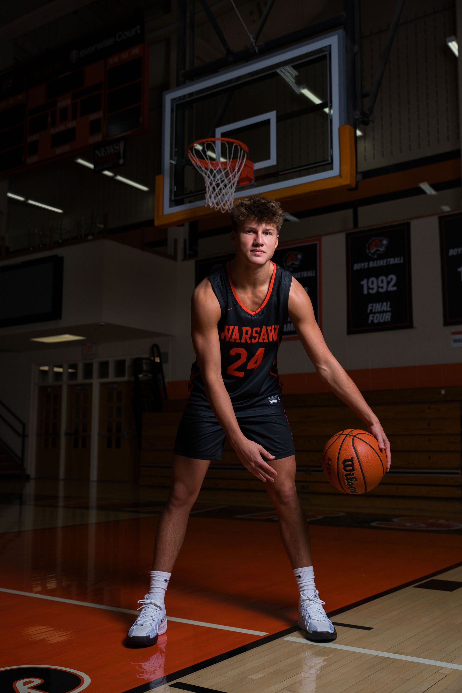 Teen basketball player in Warsaw #24 jersey holding a ball under the hoop in a school gymnasium.