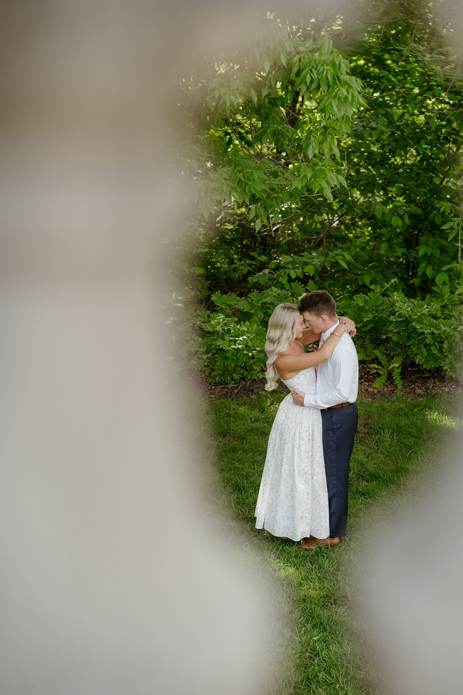 Couple embracing tenderly in a lush green garden framed by soft blurred foreground elements.