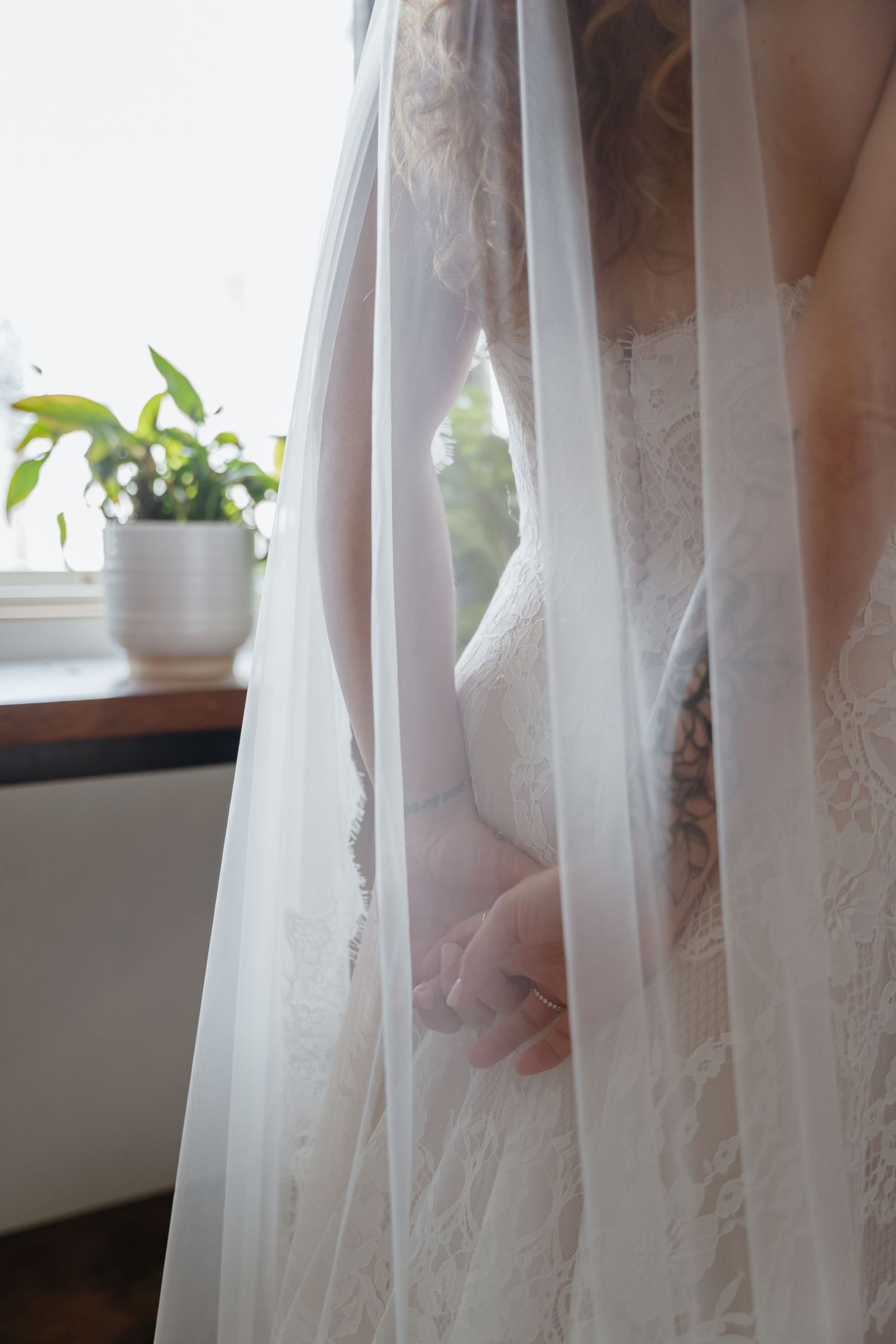 Close-up of a bride in a lace wedding gown with a flowing veil near a sunlit window.