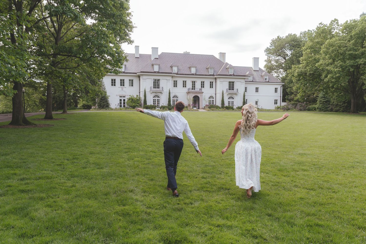 Bride and groom holding hands running toward grand white estate mansion on lush green lawn.
