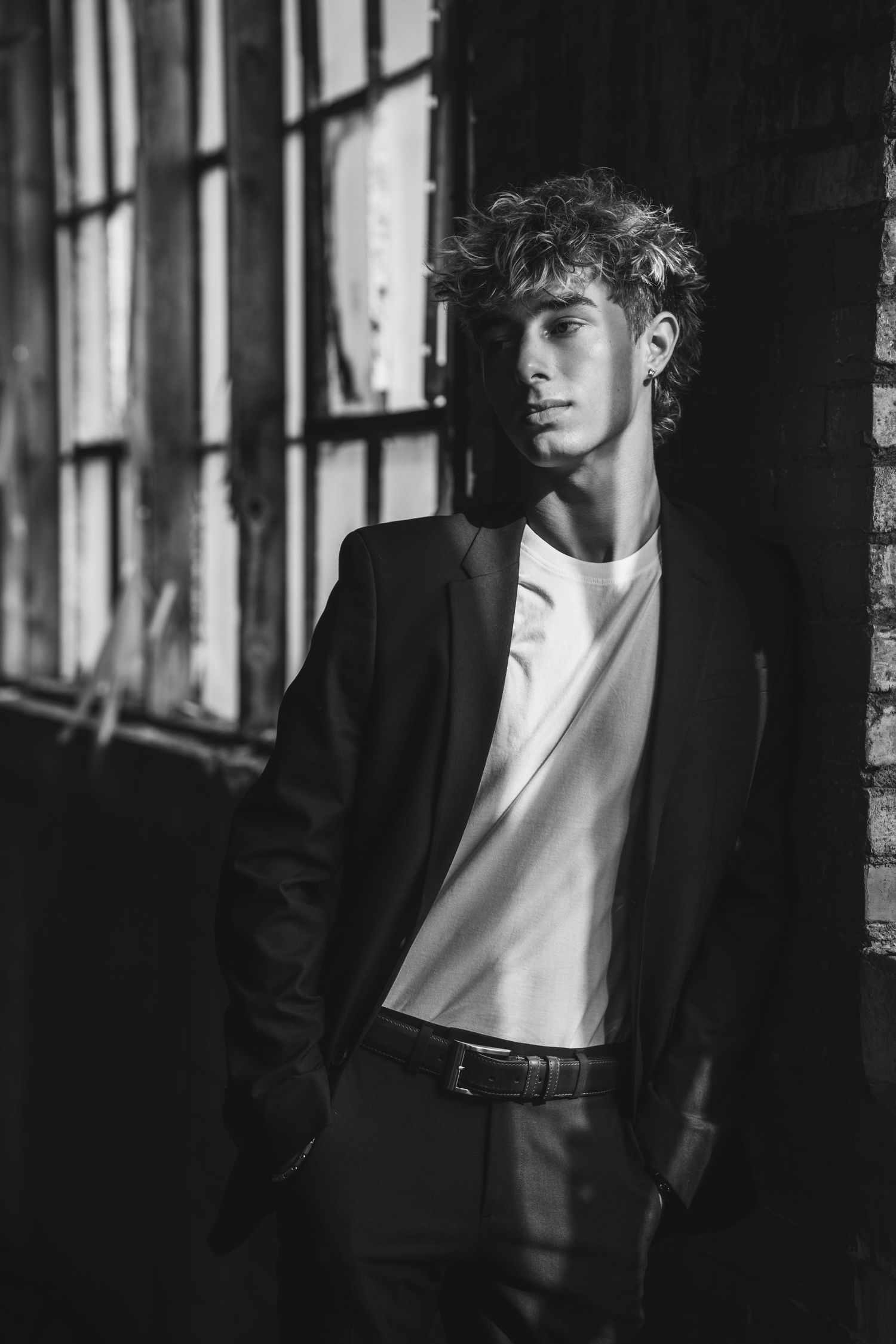 Stylish young man in black blazer and white tee poses in black and white industrial setting.