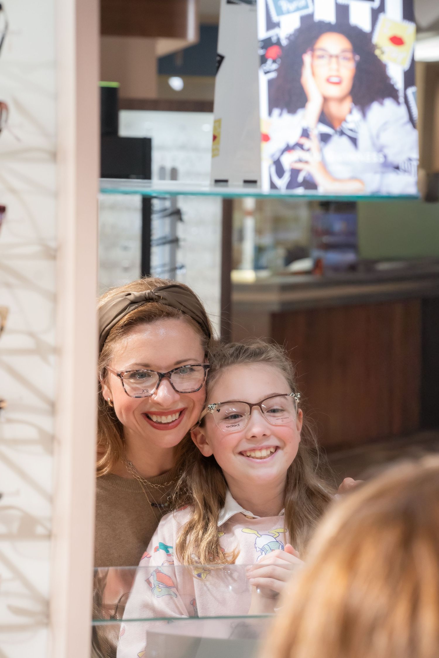 A smiling mother and daughter try on matching eyeglasses at an optical store.