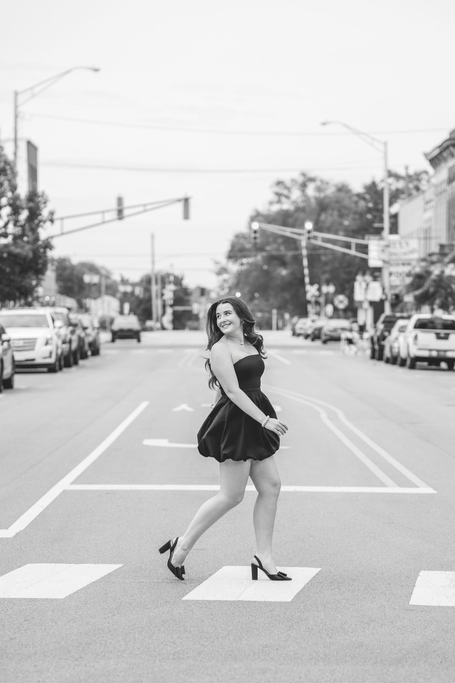 Black and white photo of young woman in black dress crossing a city street intersection.