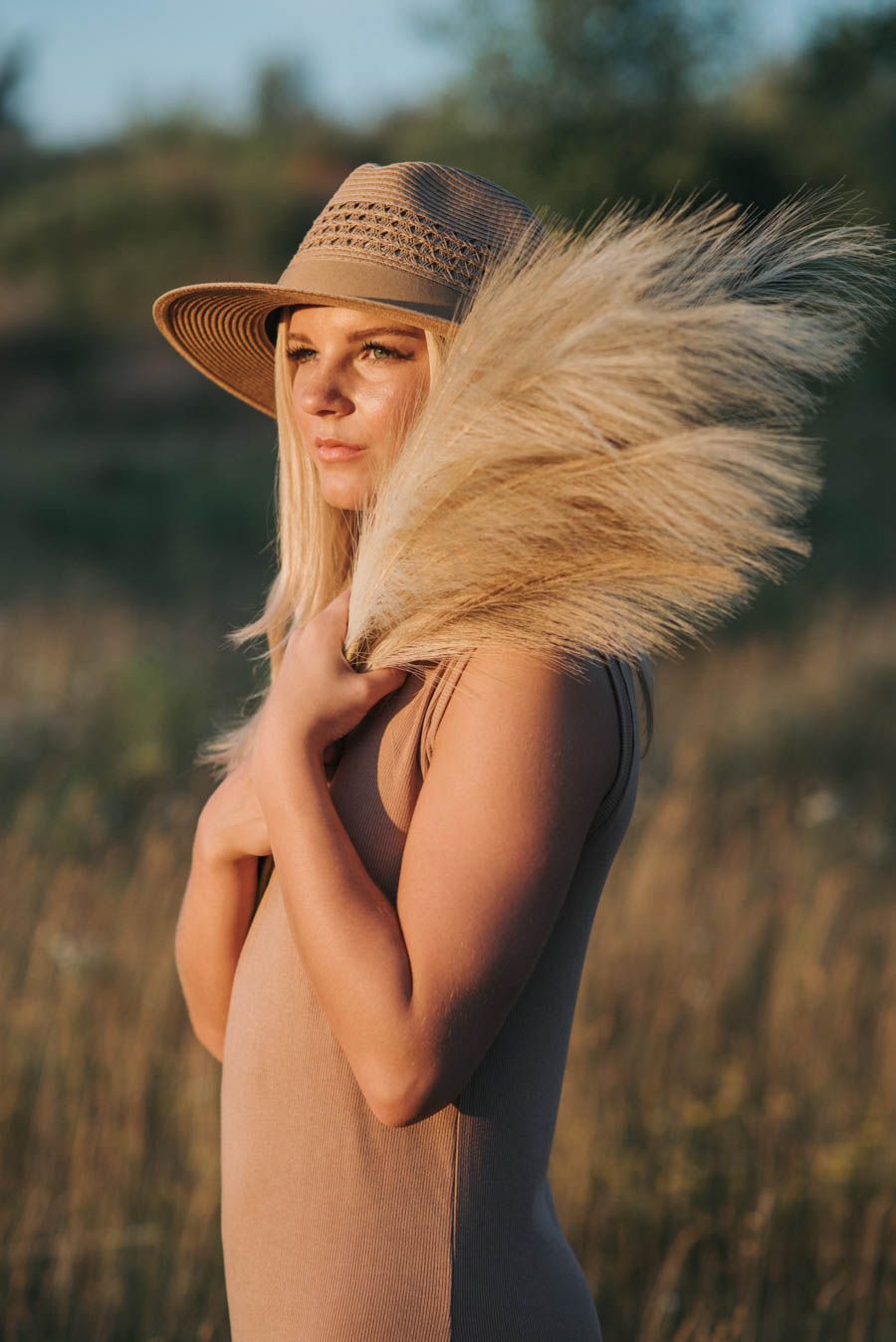 Blonde woman in straw hat holding pampas grass in a golden field at sunset.