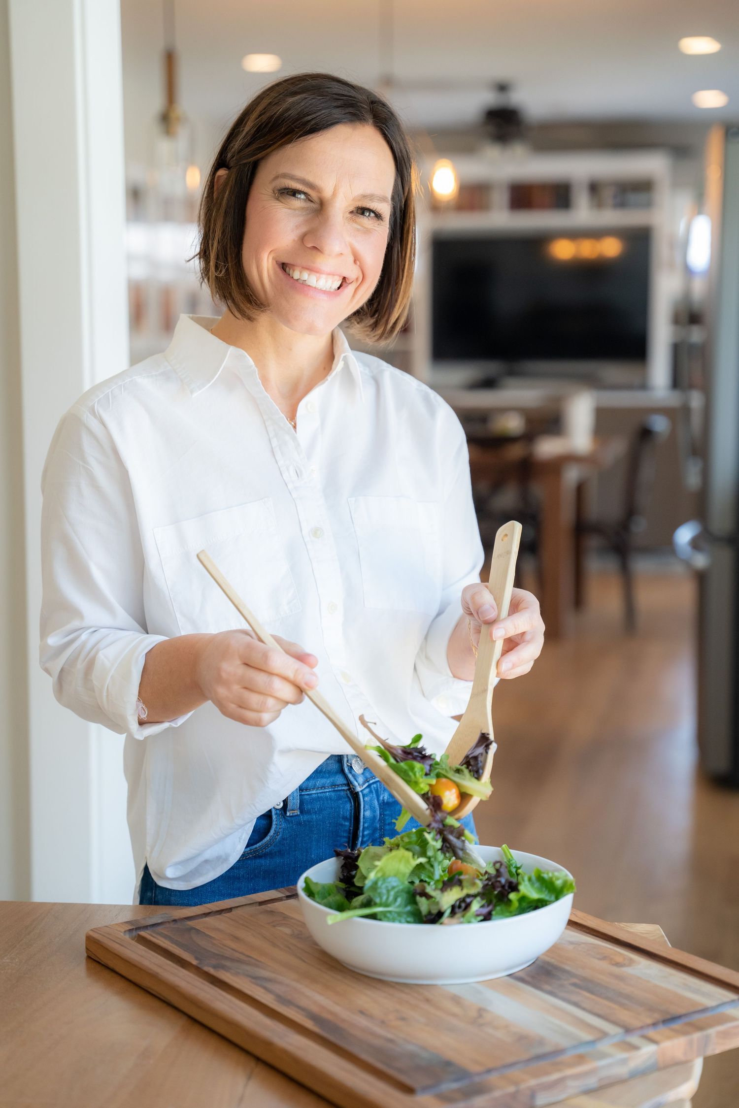 Smiling woman in white shirt tossing fresh green salad with wooden tongs in a bright modern kitchen.