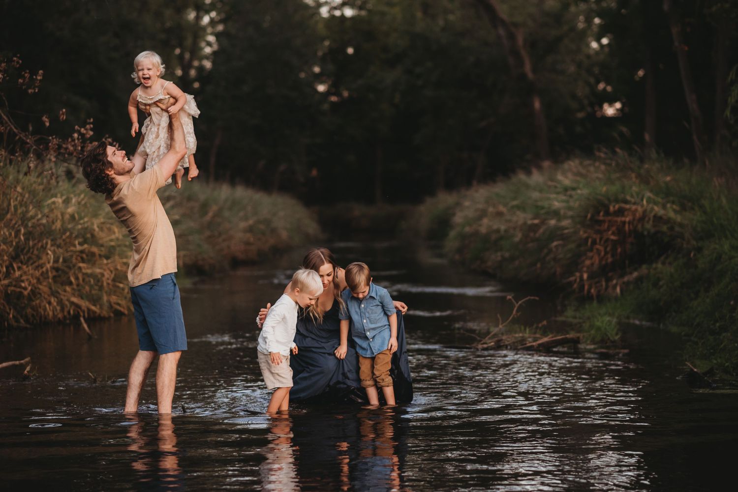 Family playing in a shallow creek surrounded by lush greenery, father lifting baby in the air.