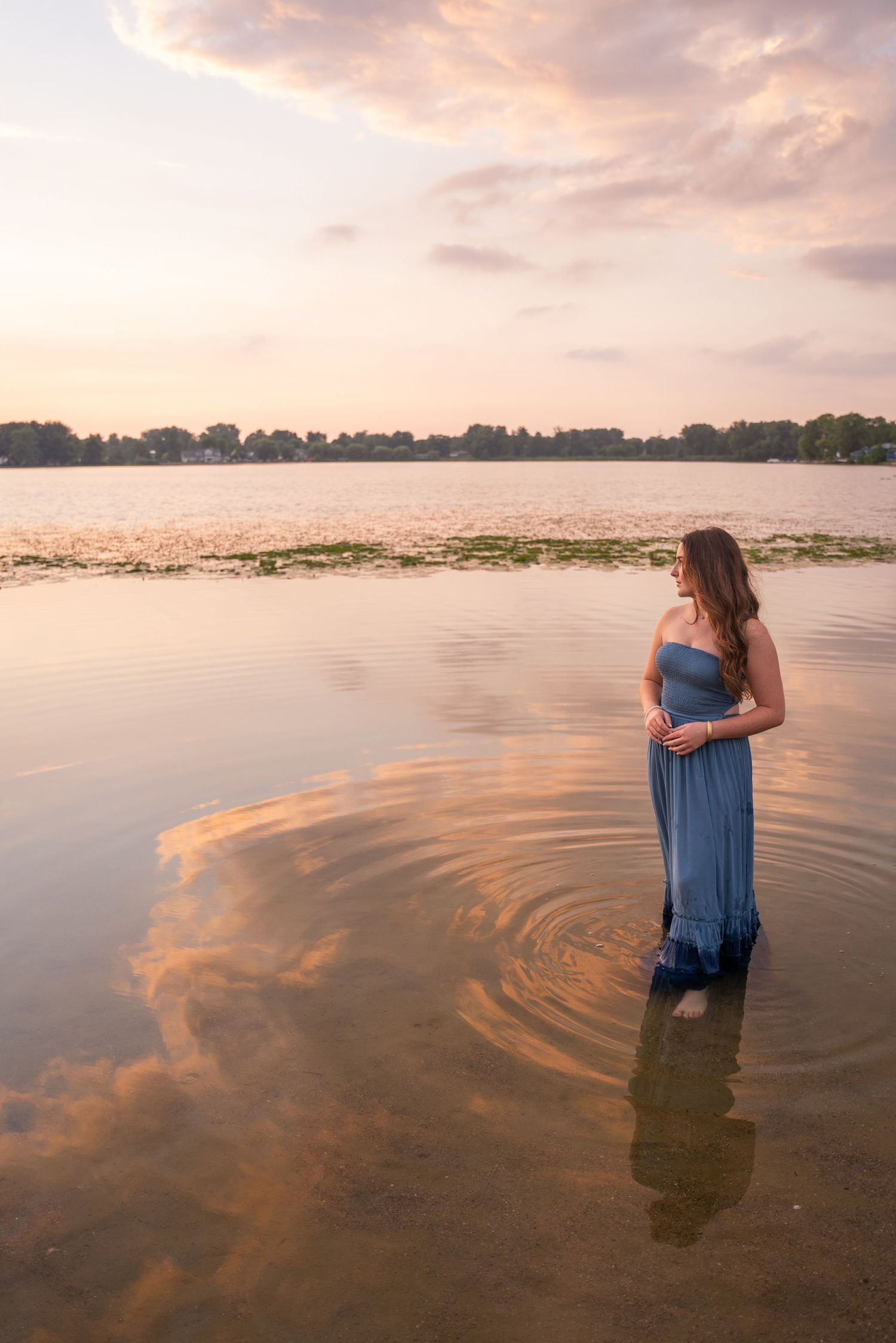 Woman in a blue strapless gown standing in a calm lake at sunset with pink clouds.