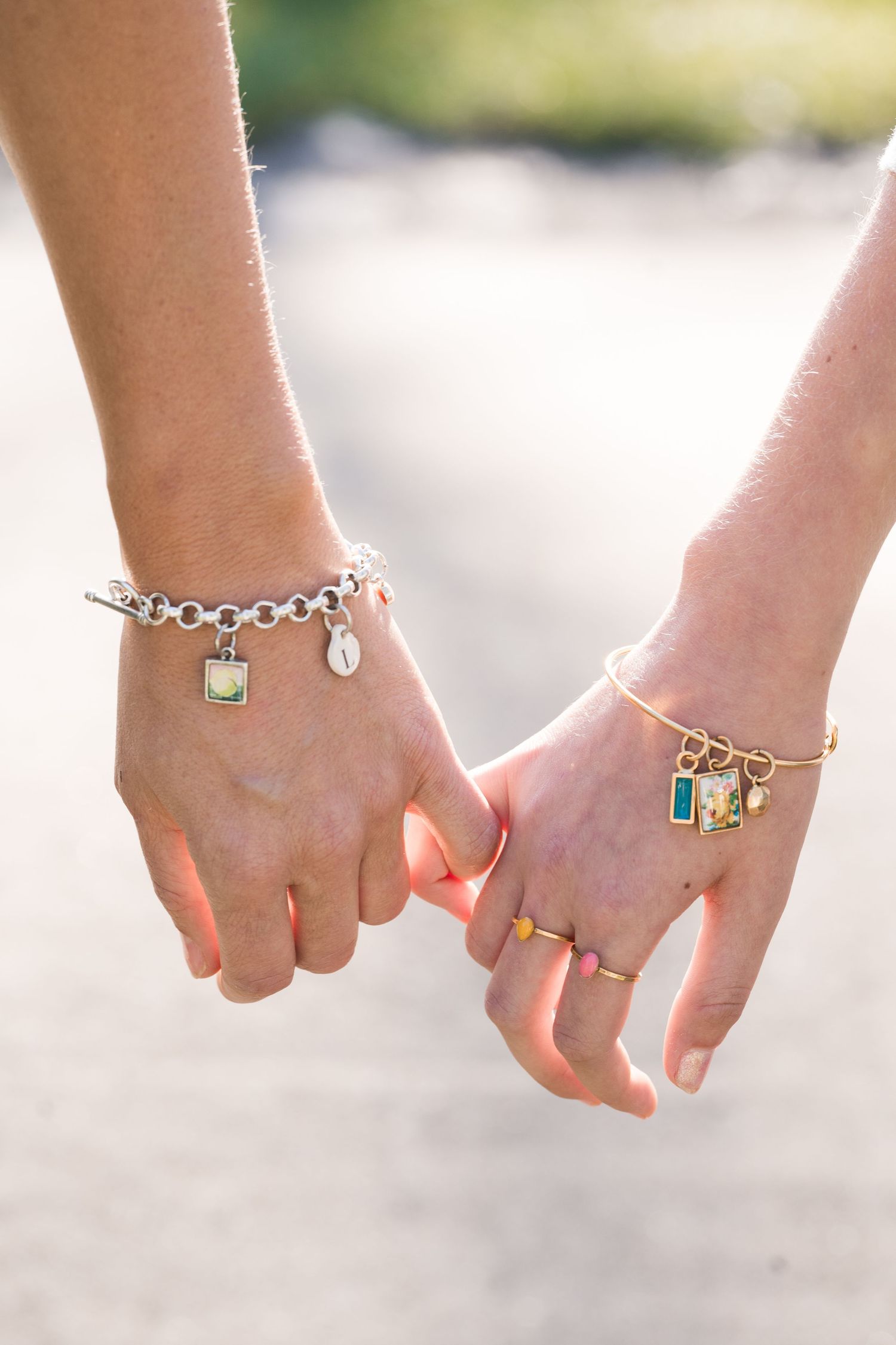 Two women holding hands showing charm bracelets and rings in outdoor sunlight.