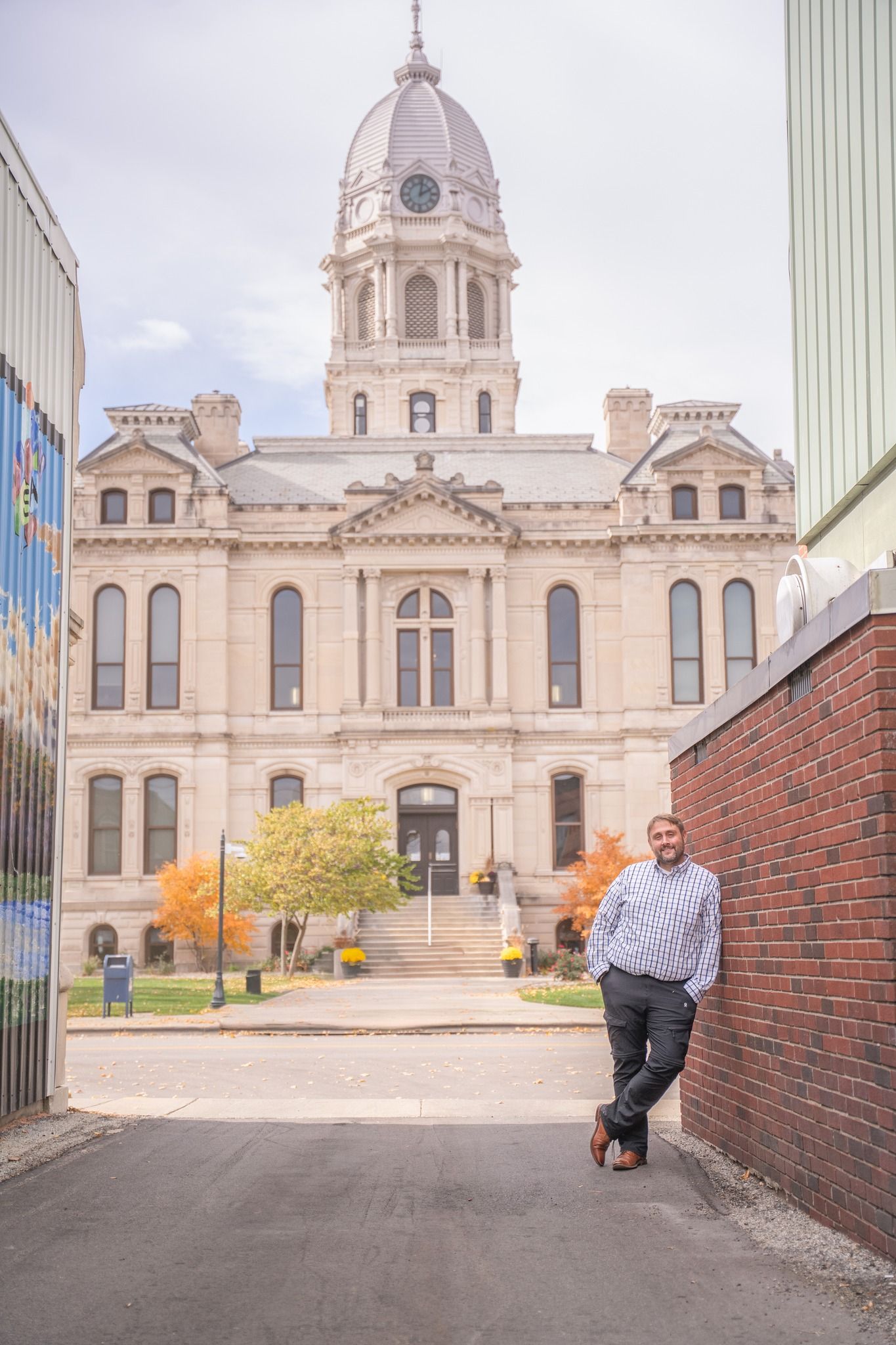 Man leaning against brick wall with historic domed courthouse and autumn trees in the background.