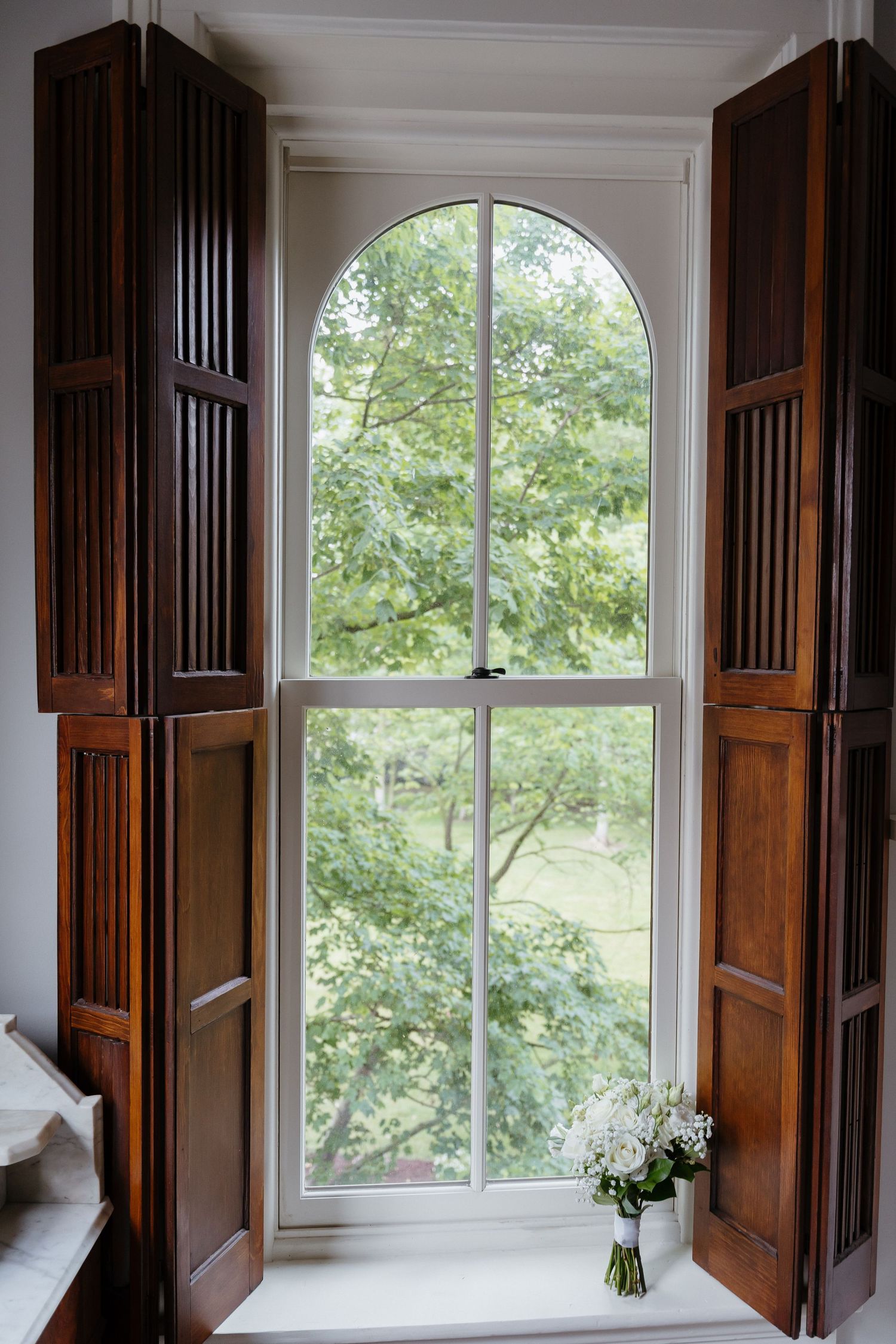Elegant arched window with wooden shutters and bridal bouquet on windowsill overlooking garden.