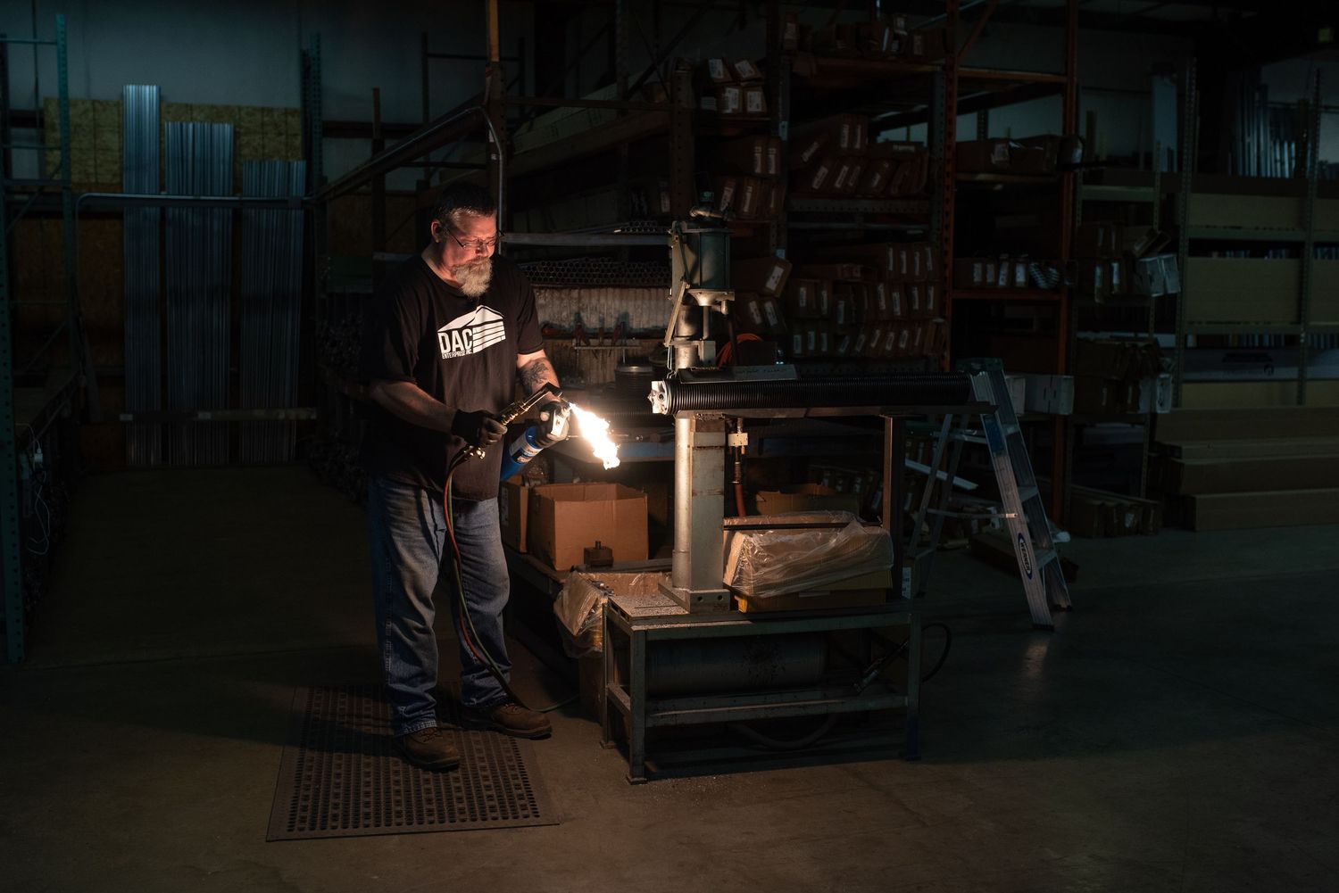 Worker using a bright flashlight to inspect machinery in a dark industrial warehouse at night.