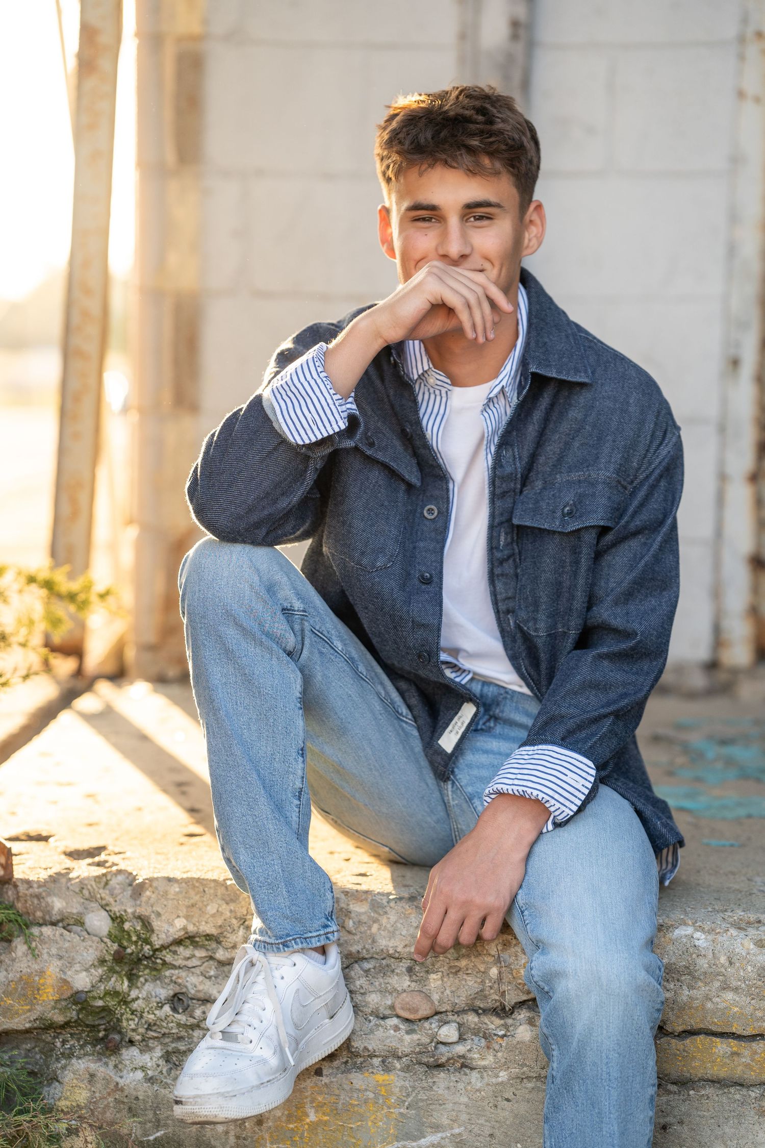 Young man in denim jacket and jeans sitting on steps outdoors in golden sunlight.