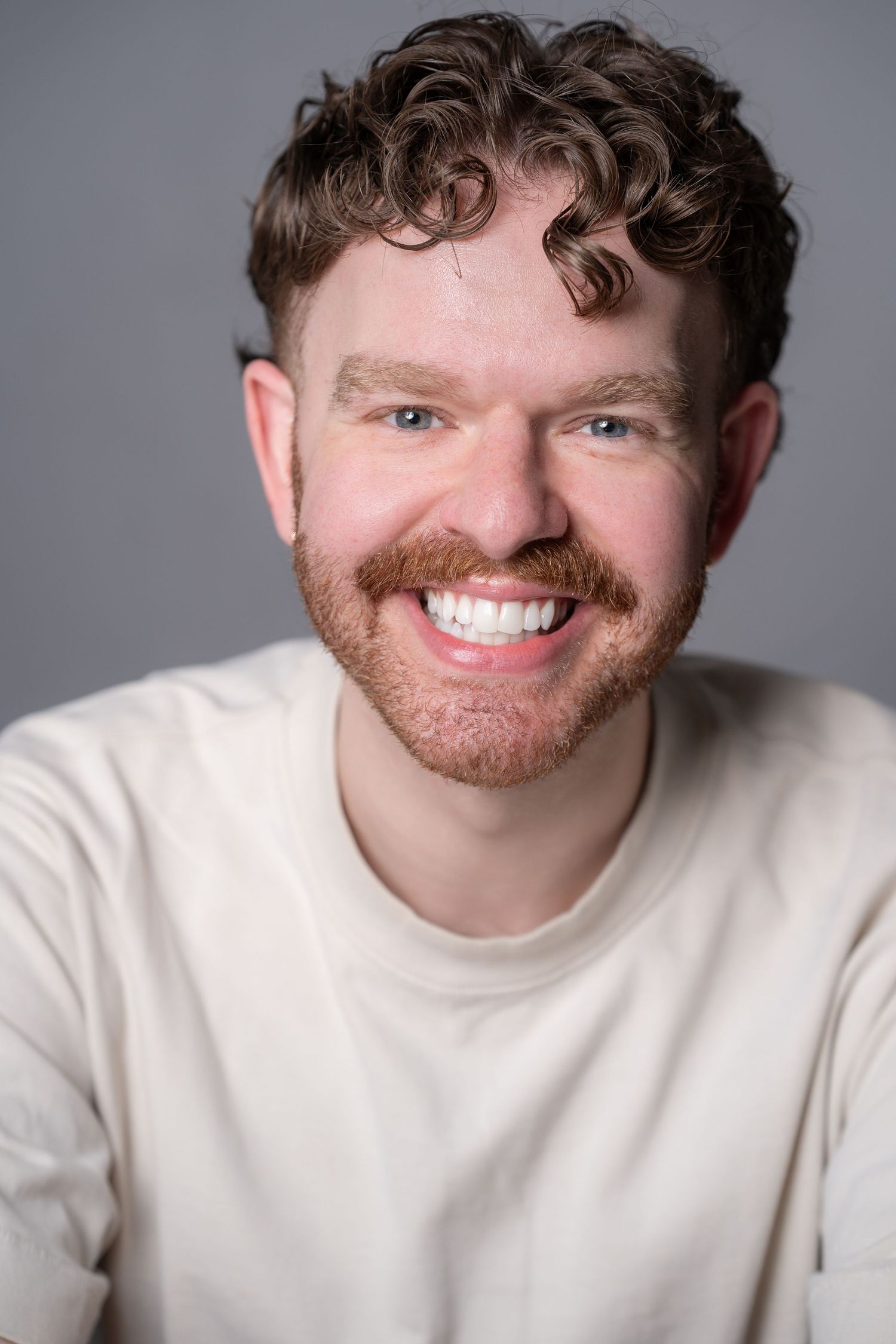 Young man with curly hair and a mustache smiling confidently against a gray background.