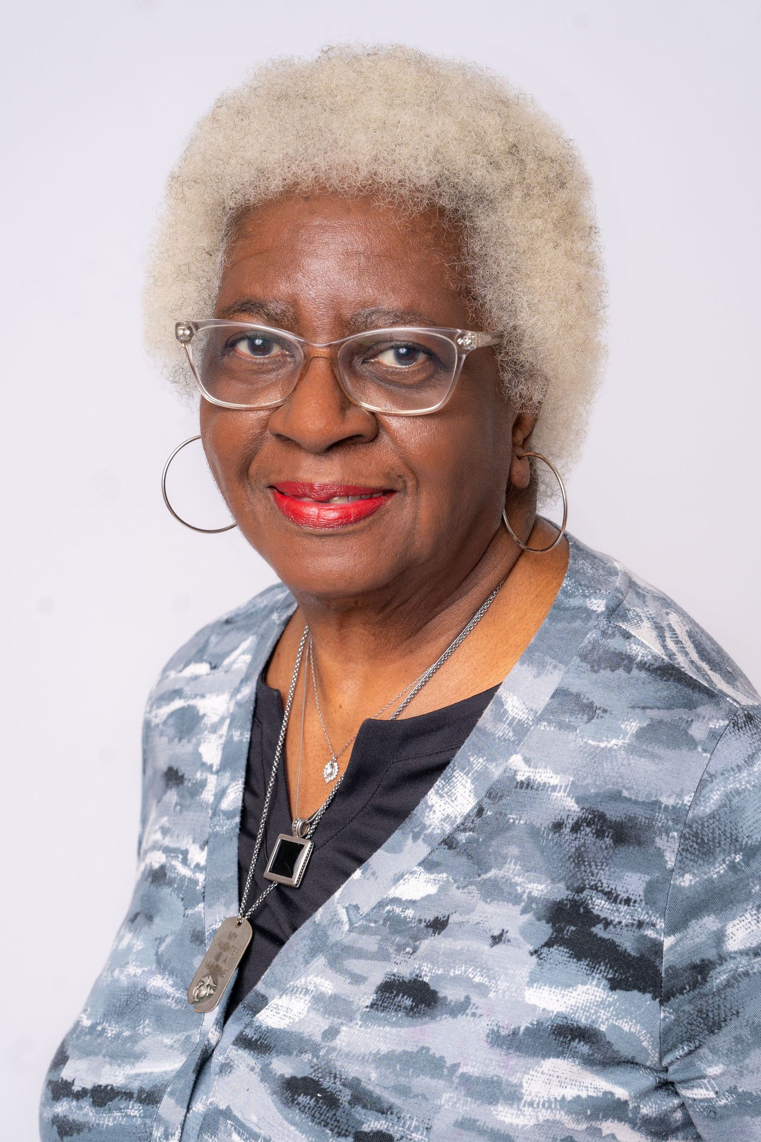 Elderly Black woman with white hair, glasses, and hoop earrings smiling in a gray patterned blazer.
