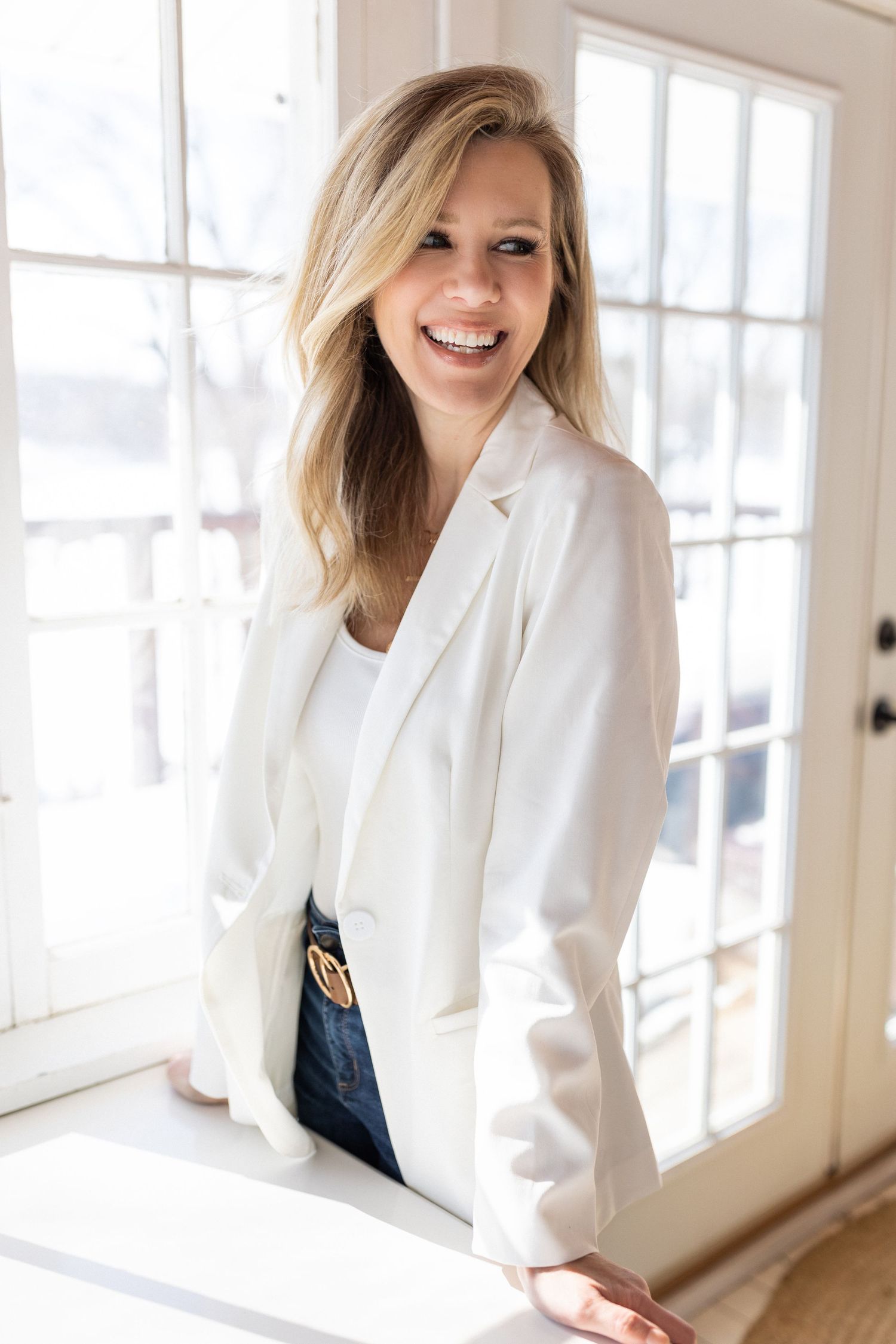 Laughing blonde woman in white blazer leaning against French doors in bright sunlit room.