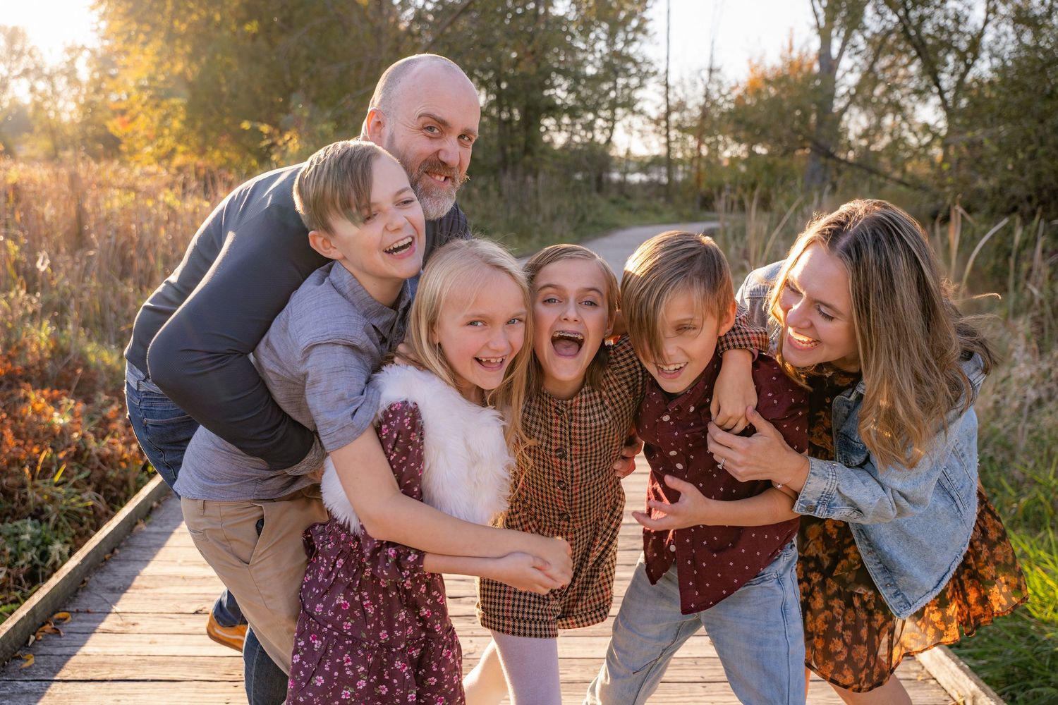 A joyful family of six hugging and laughing together on a wooden bridge during golden hour.
