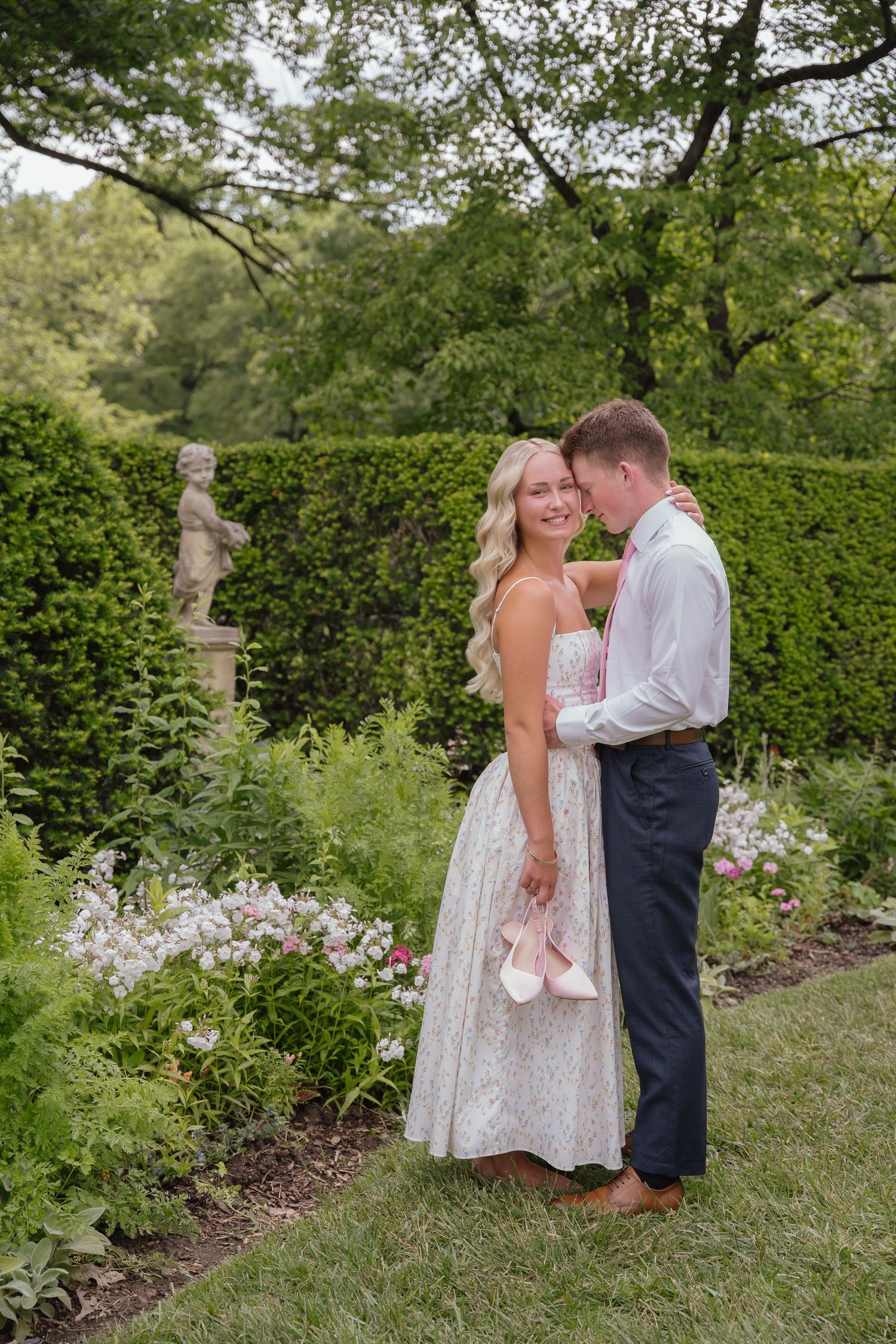 Couple embracing in a lush garden, woman in white lace dress holding pink shoes, man in white shirt and navy pants.