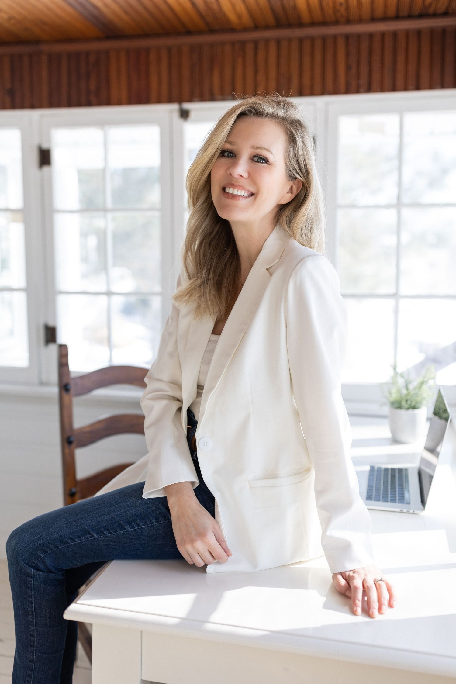 Happy blonde woman wearing white blazer and dark jeans seated on white desk indoors.