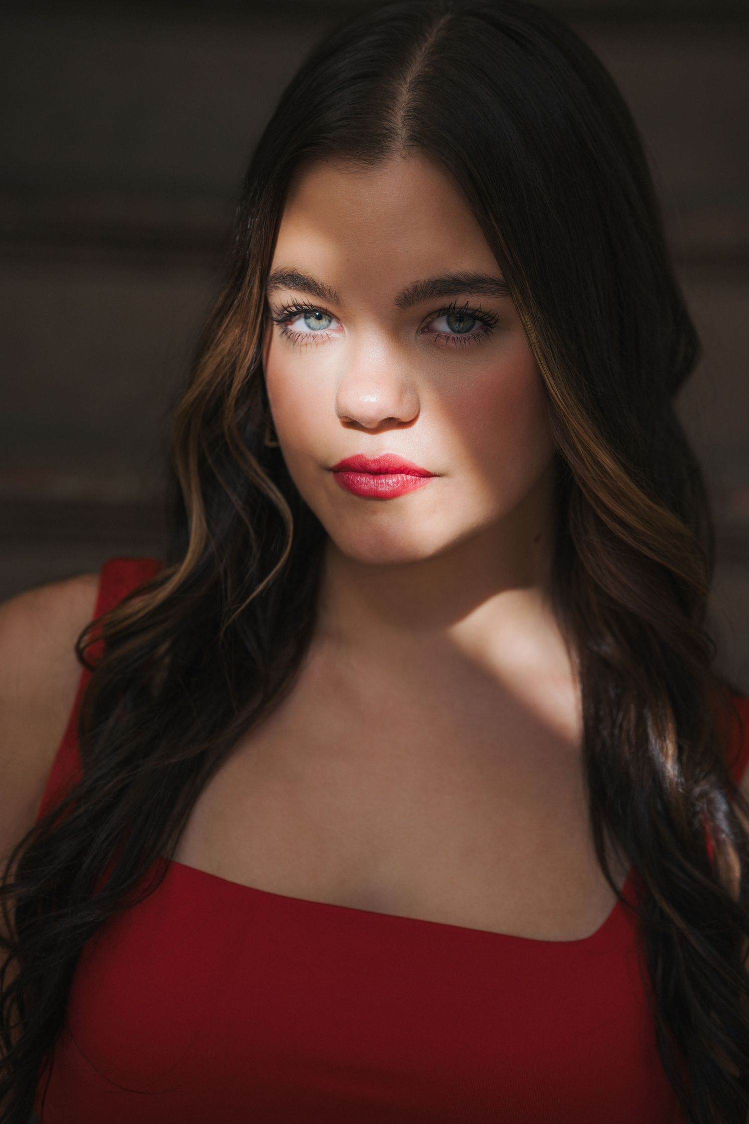 Brunette woman with blue eyes and red lips wearing red top in dramatic shadow lighting portrait.