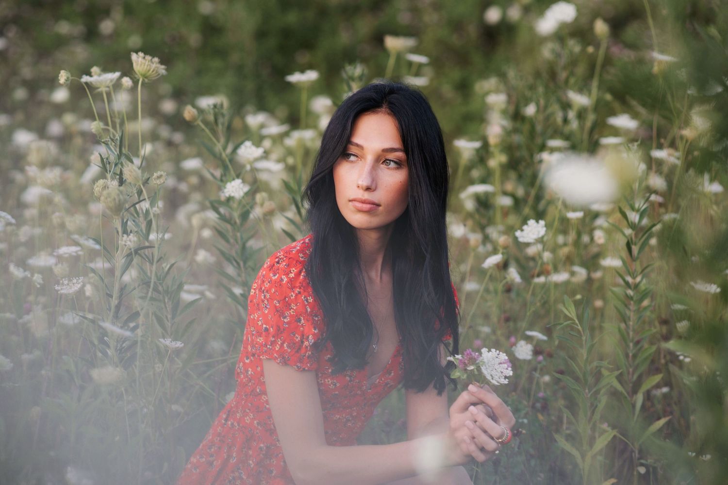 Young woman in red floral dress sitting among white wildflowers in a field.