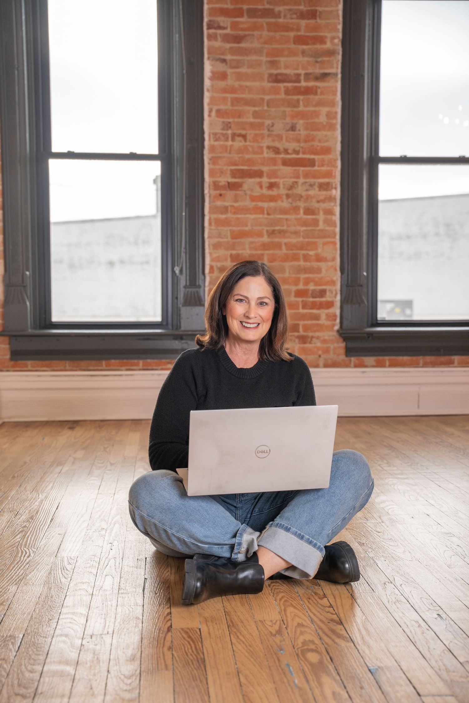 A smiling woman sits cross-legged on a hardwood floor using a Dell laptop.