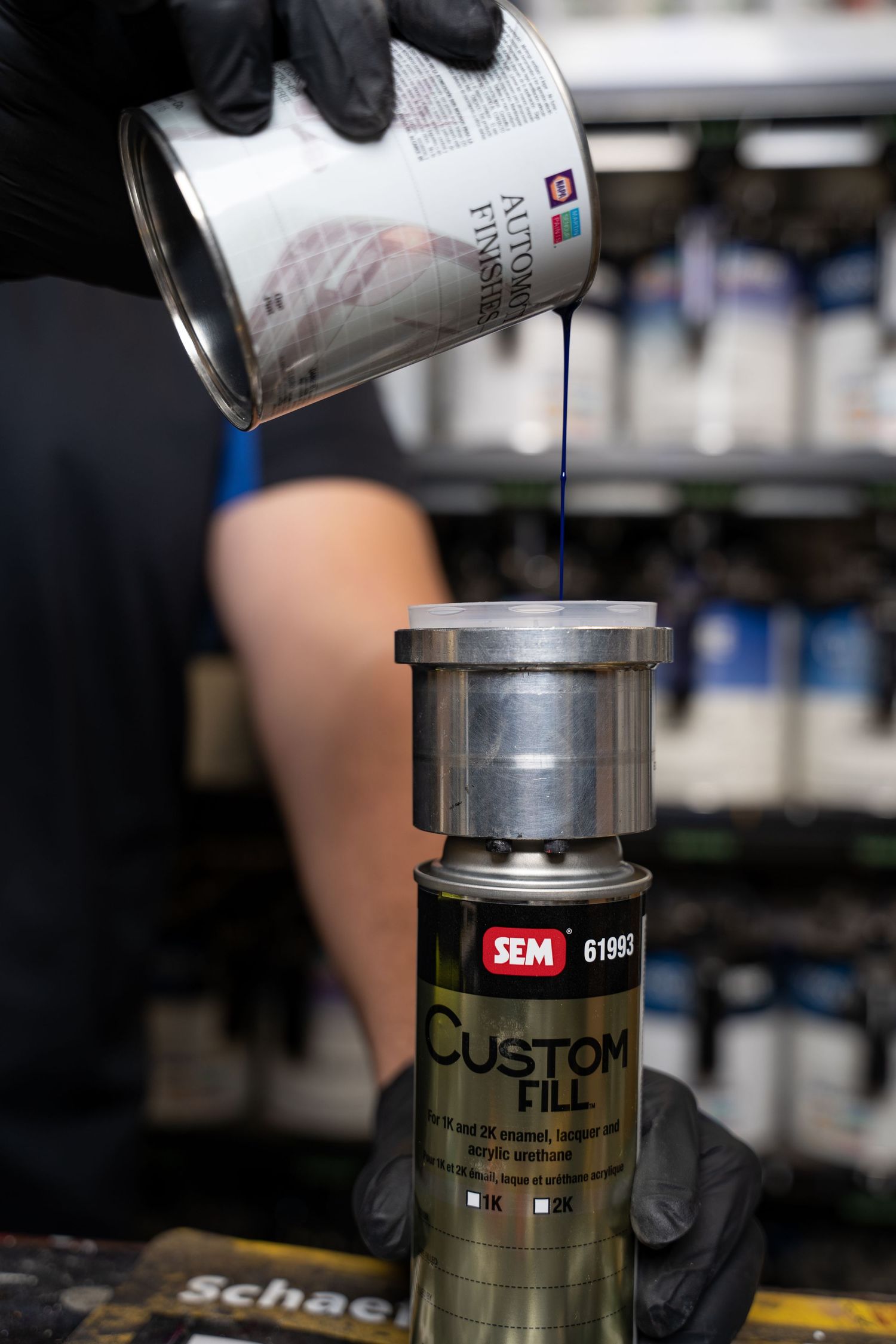 Gloved hands pouring purple automotive paint into a SEM Custom Fill canister in an auto shop.