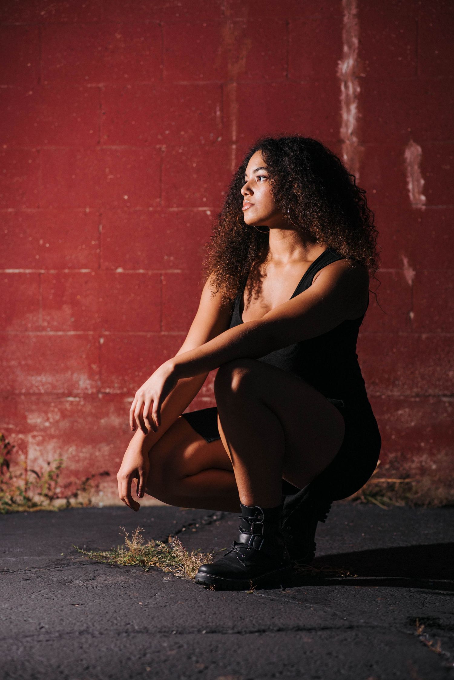 Young Black woman with curly hair crouching in black outfit and boots against a red brick wall.