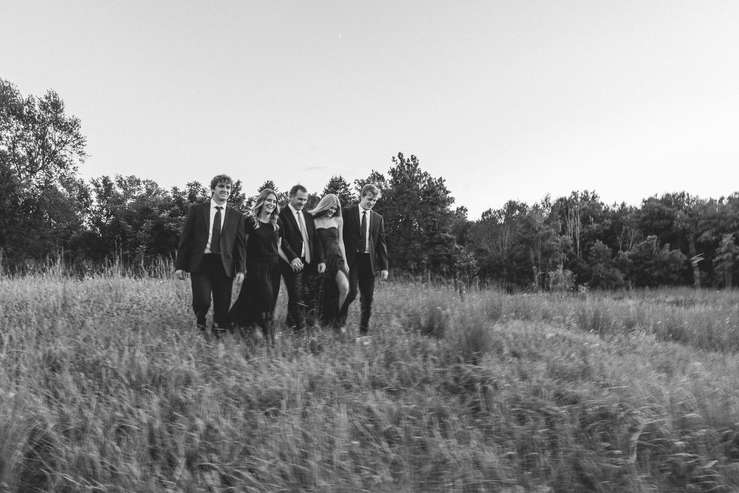 A black and white photo of five people walking together through a tall grass field at dusk.