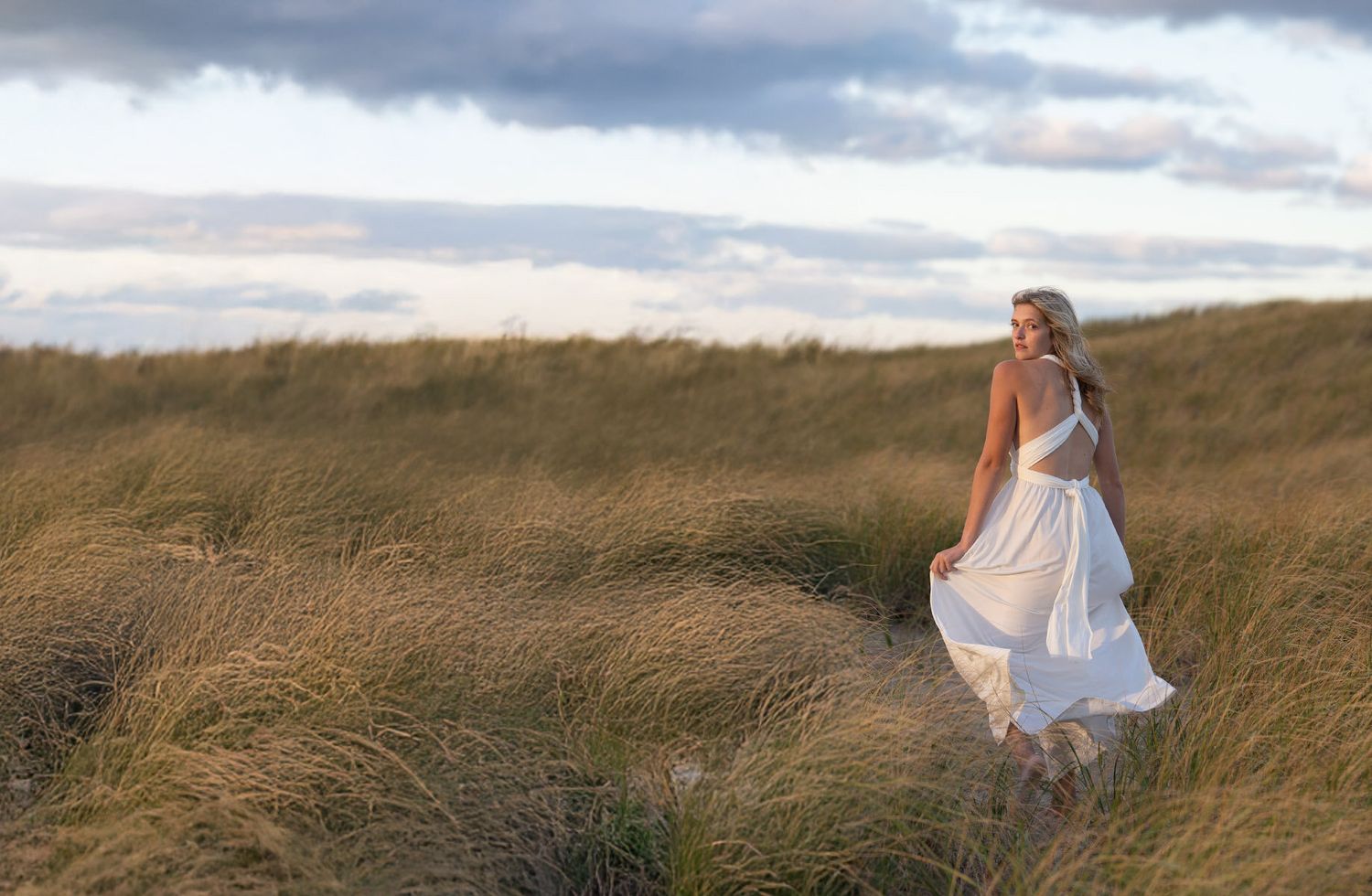 Girl in white dress walking through golden coastal grass dunes at sunset.