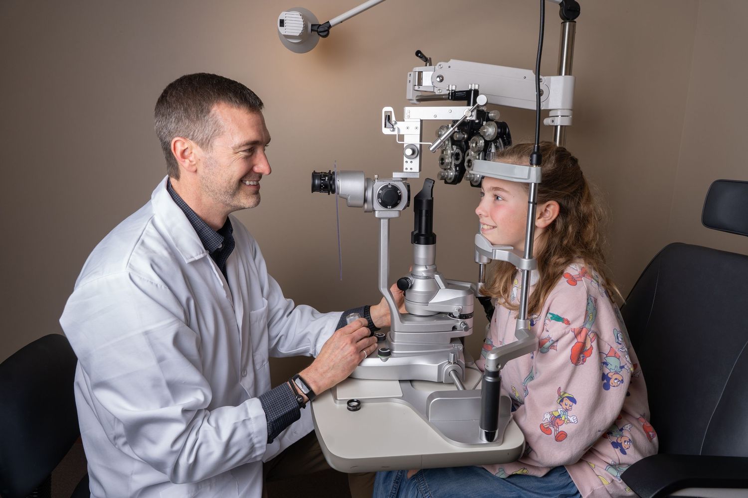 Male optometrist examining young girl's eyes using slit lamp in modern eye clinic.
