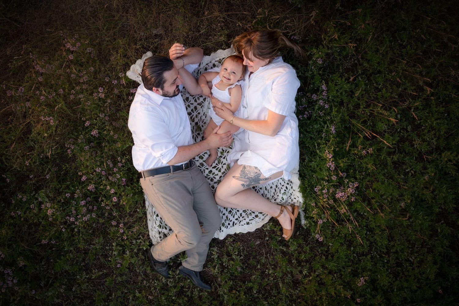 Aerial view of two parents lying on grass with their baby between them in a serene outdoor setting.