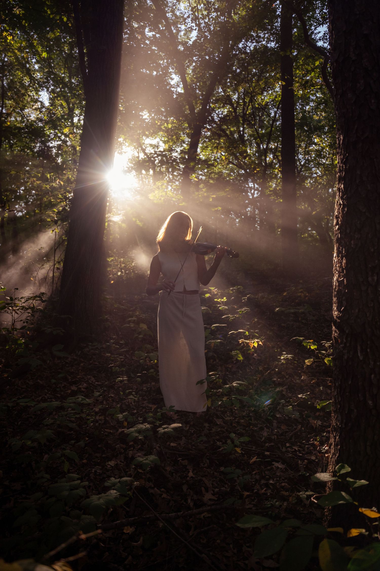 Woman in white dress playing violin in a misty forest with golden sunlight rays streaming through trees.