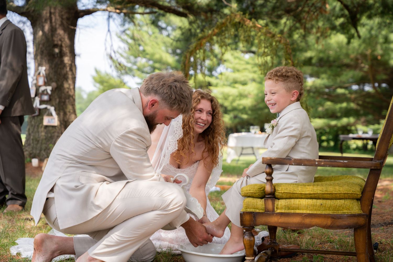 Groom washing feet of bride and smiling child during an outdoor wedding ceremony ritual.