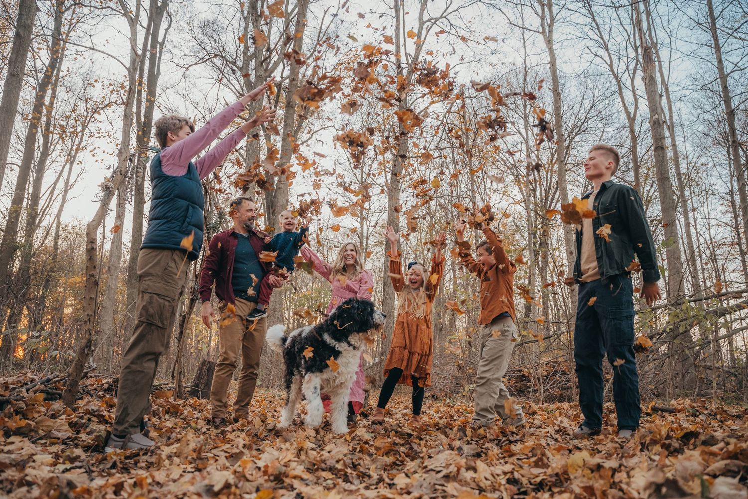 Family with dog tossing autumn leaves in a bare winter forest during a fun outdoor photo session.