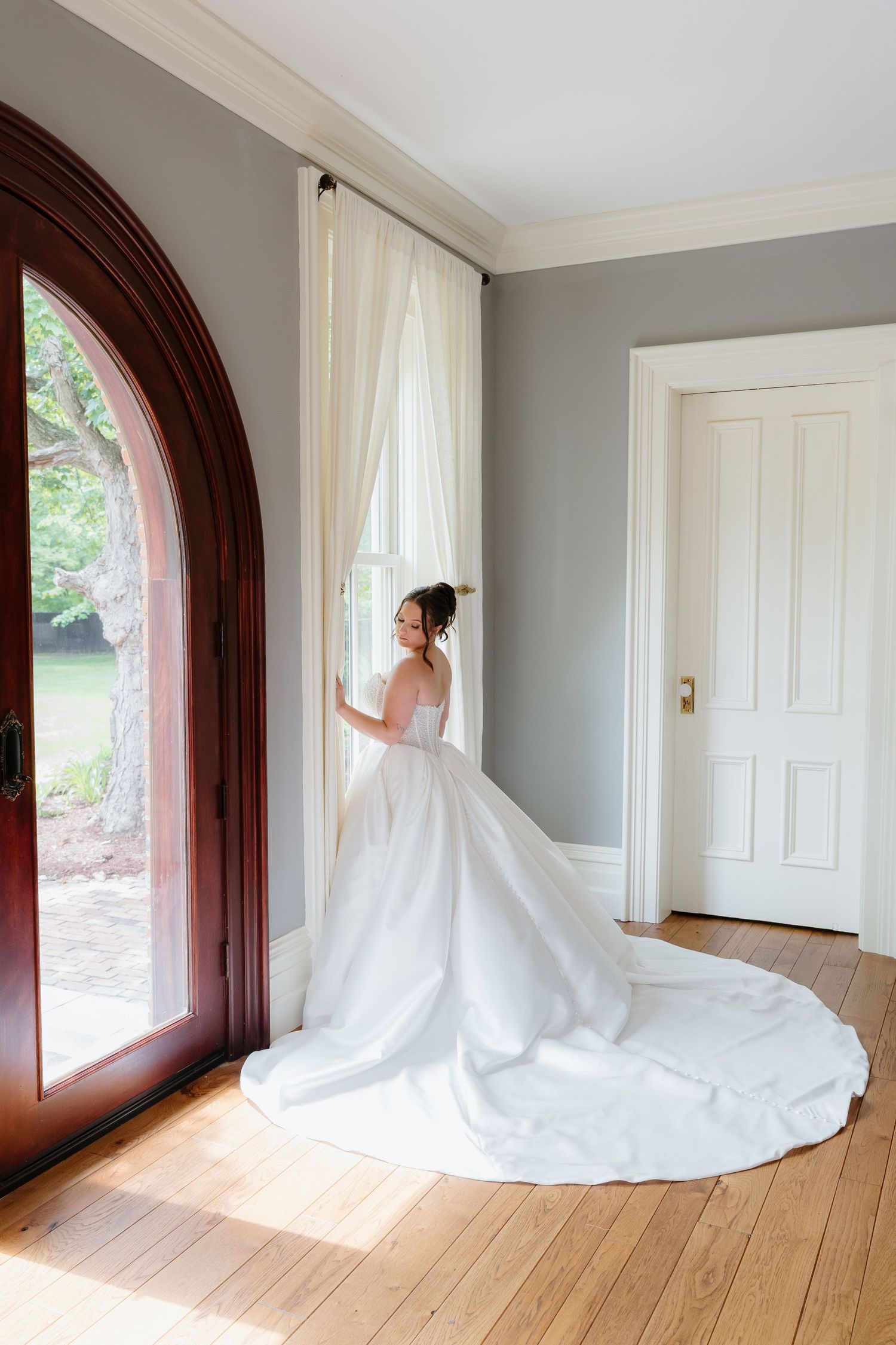 Bride in a white ball gown with long train standing by a tall arched window in a classic room.