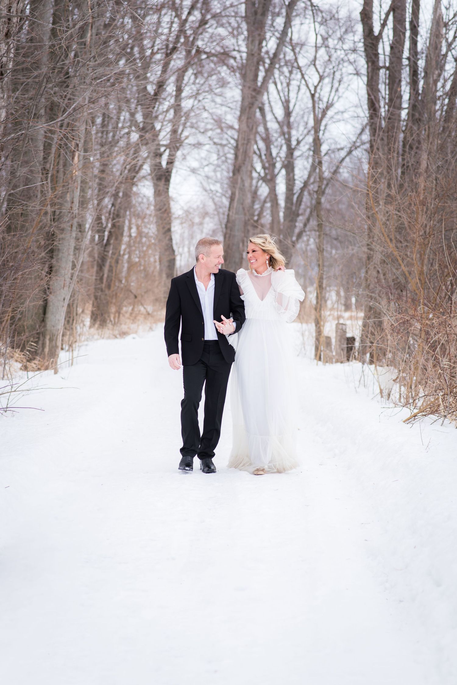 Bride and groom walking hand in hand down a snowy winter path surrounded by bare trees.