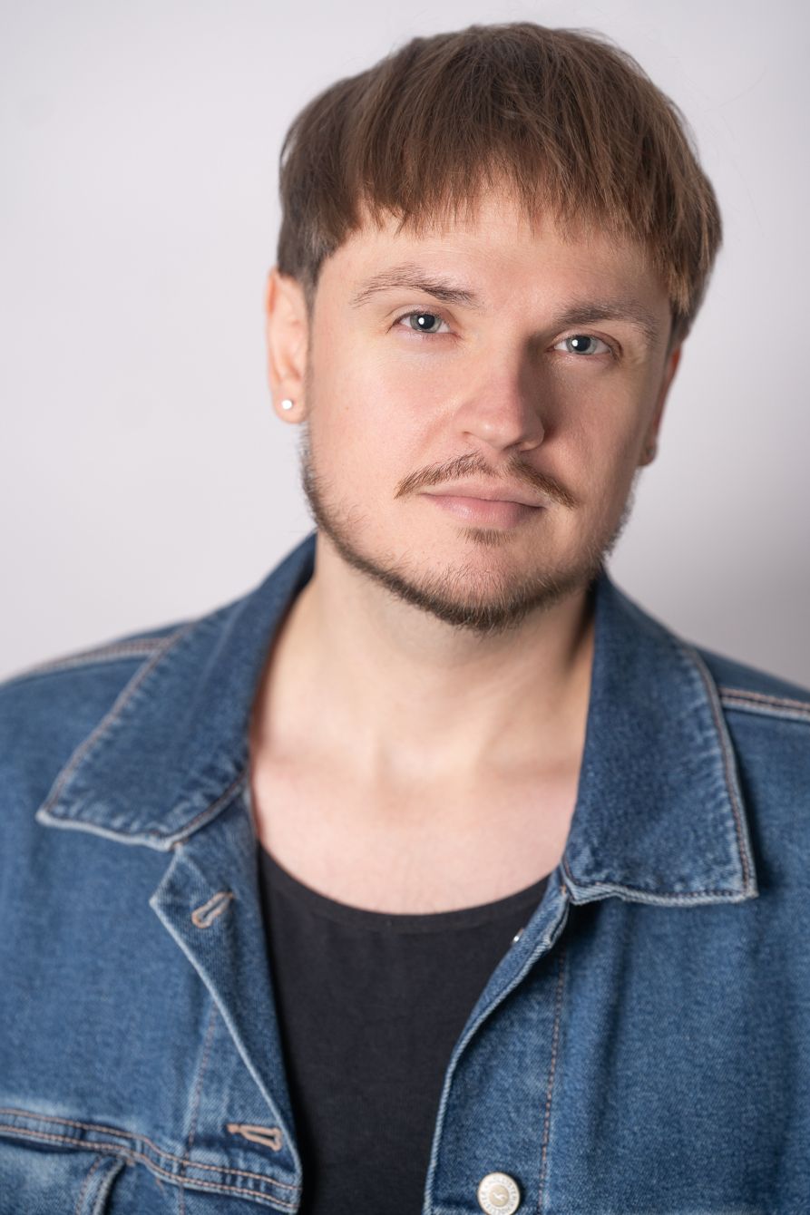 Young man with light brown hair and stubble wearing a denim jacket against a white background.