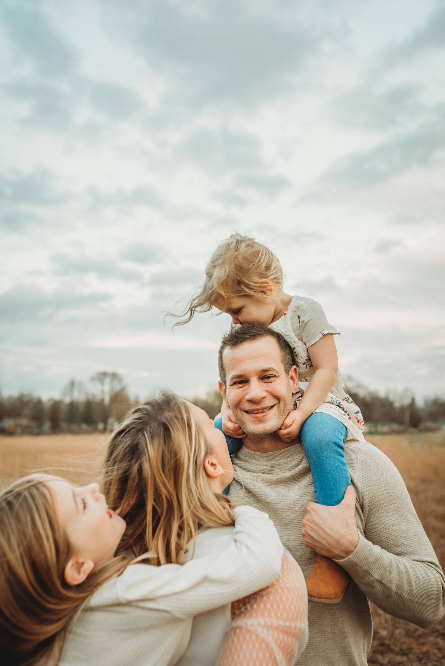 Happy father carries toddler on shoulders while mother embraces them in open field.
