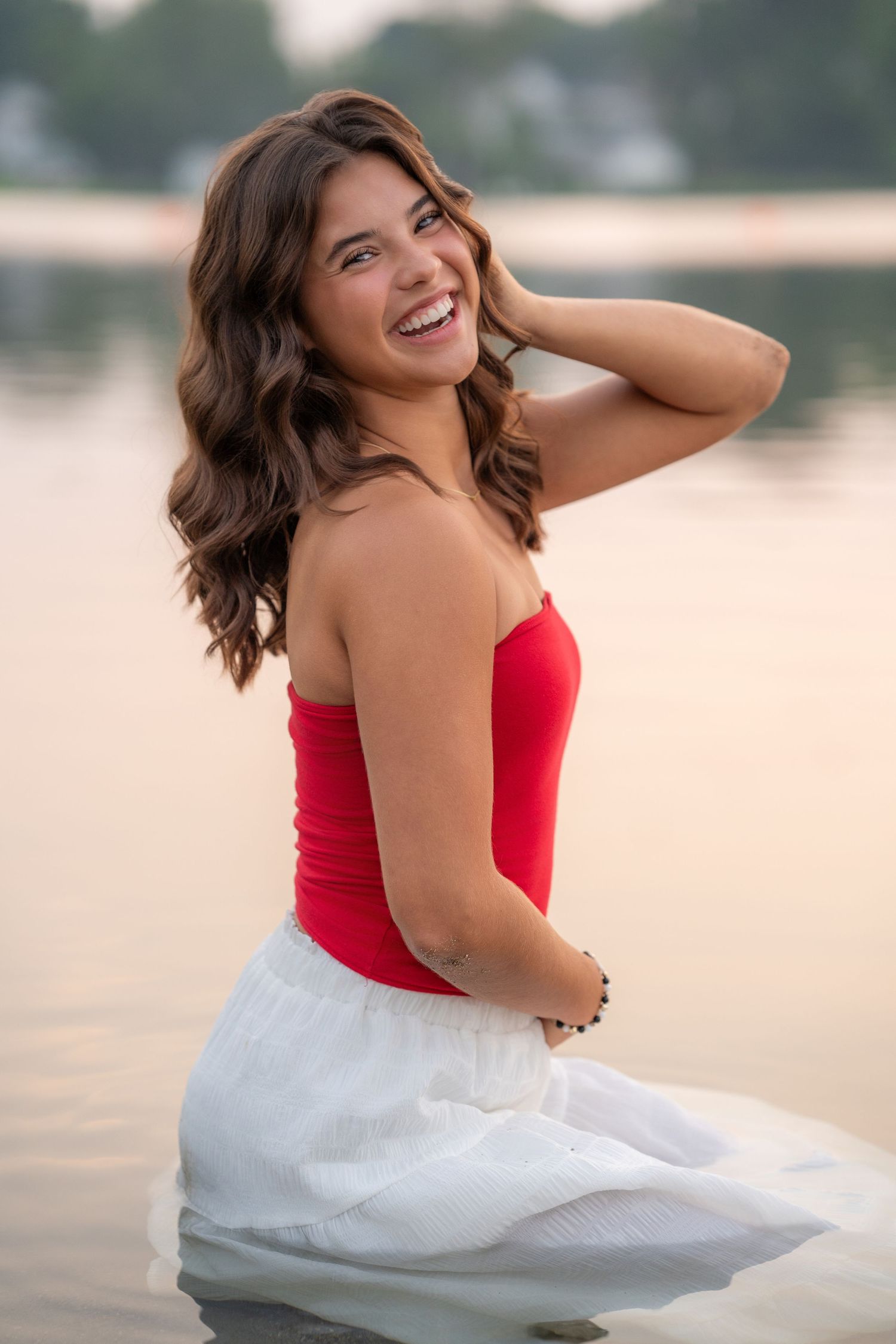 Smiling young woman in red top and white skirt posing by a lake at sunset.